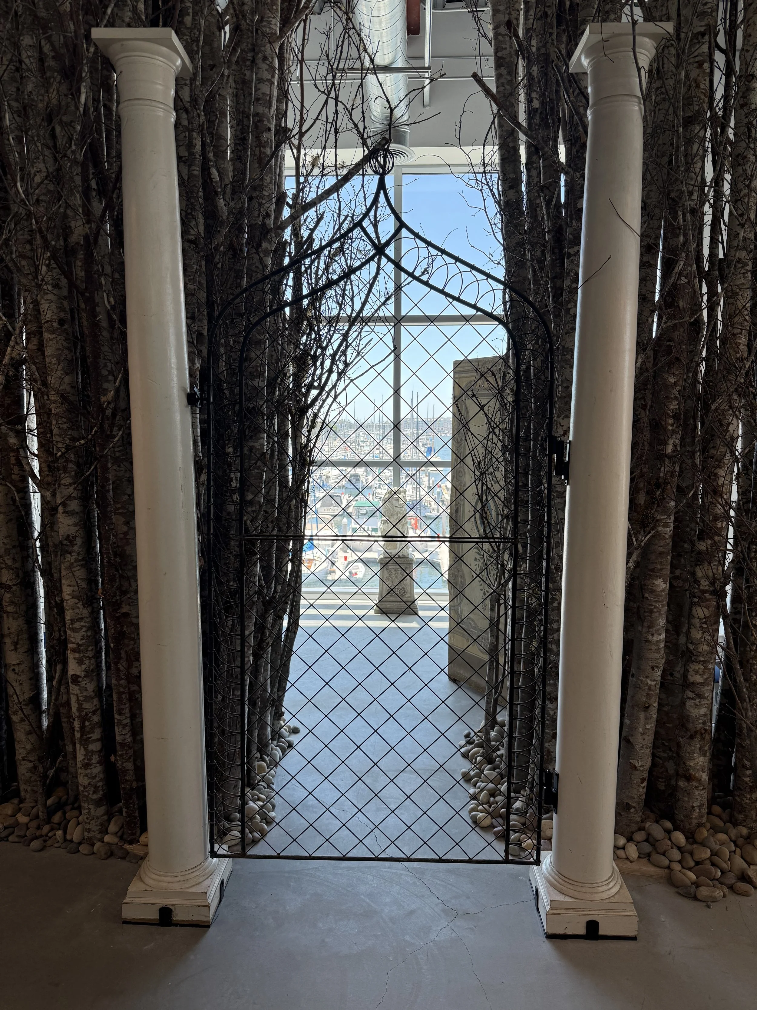 A decorative metal gate with a pointed arch, flanked by two white columns, leading to a balcony with a view of a marina and city skyline in the background.
