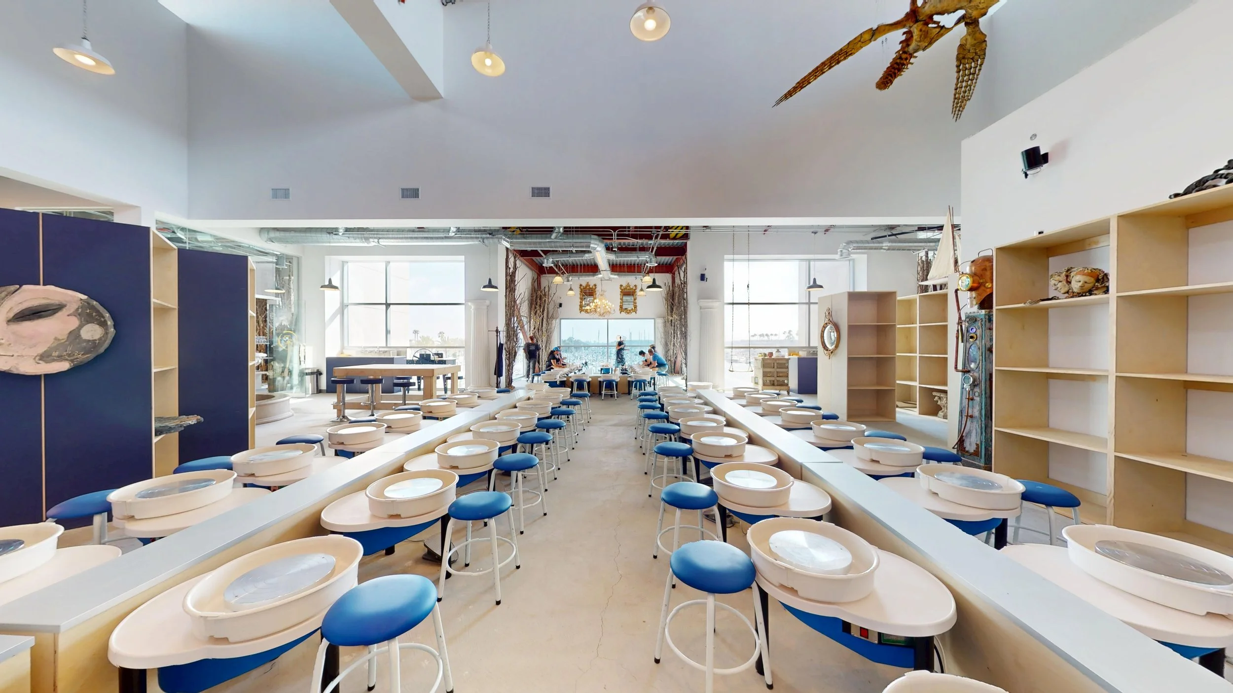 Empty pottery studio with circular pottery wheels and blue stools, large windows, and artistic decorations.