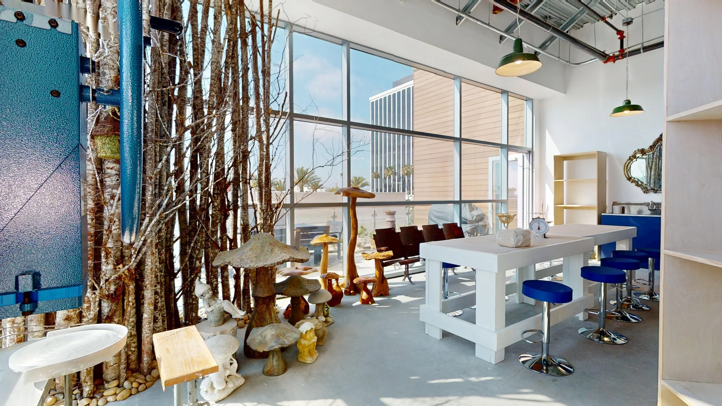 Interior space with large floor-to-ceiling windows, decorated with artificial trees and mushroom sculptures, high tables with blue stools, and a kitchen area with shelves.