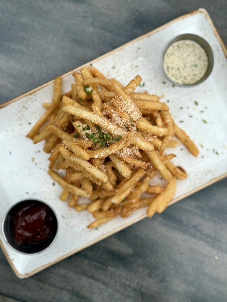 Pile of crispy French fries garnished with chopped green herbs, served with ketchup and a creamy dipping sauce on a rectangular white plate.