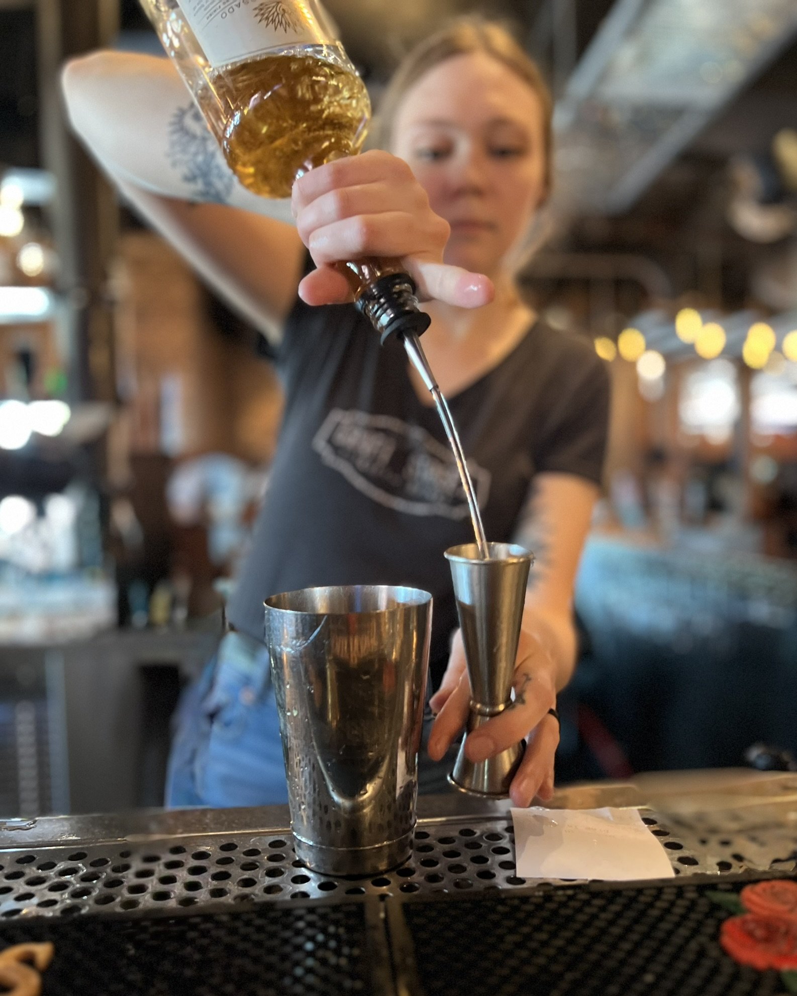 A bartender pouring a drink through a metal jigger into a shaker at a bar, with a blurred background and warm lighting.
