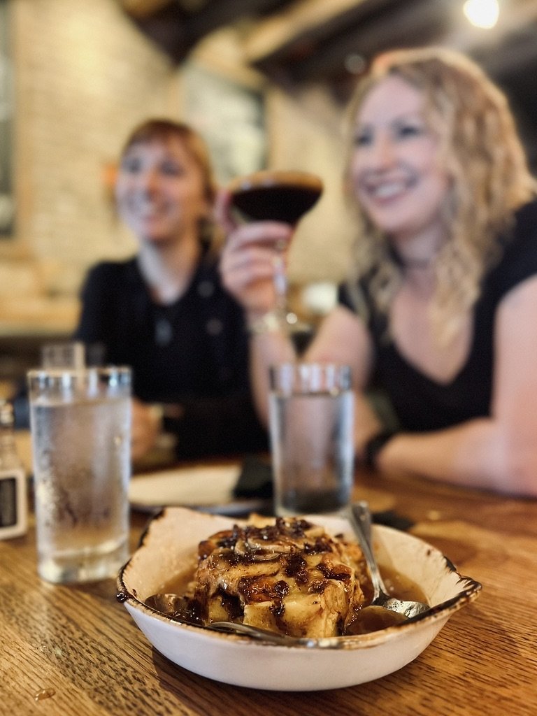 A dessert in a bowl on a wooden table in a restaurant, with two women smiling and one holding a cocktail in the background.