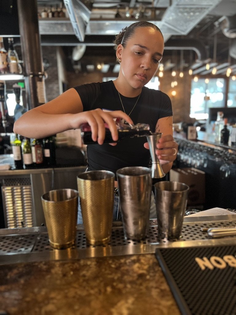 A bartender pours a drink into a metal cup at a bar with four other similar cups lined up on the counter. The bar has a lively atmosphere with bottles and bottles of alcohol in the background.