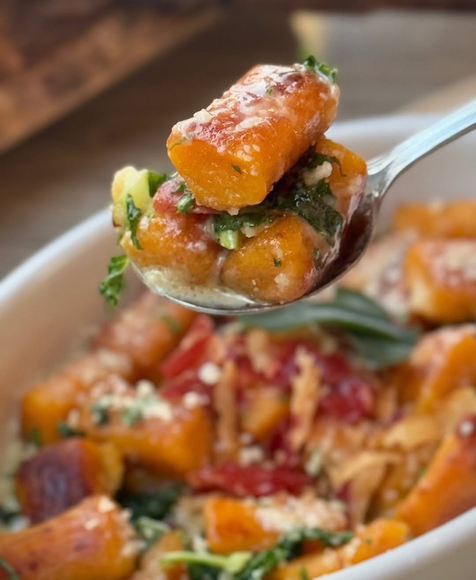 Close-up of a spoonful of sweet potato casserole with brown sugar, marshmallows, and green herbs, with more casserole in the background.