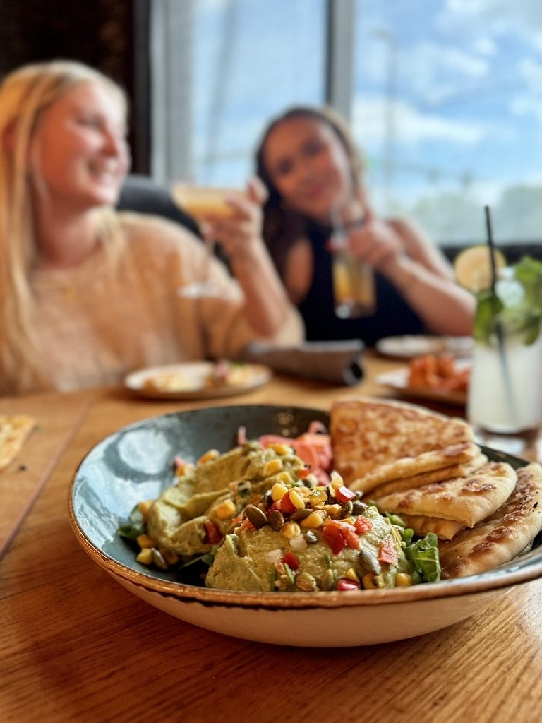 A bowl of guacamole with toppings and quesadillas on a wooden table, with two women in the background enjoying drinks at a restaurant.