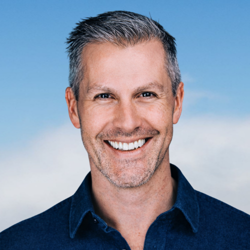 Headshot of a smiling man with short gray hair, blue eyes, and light skin, wearing a dark collared shirt against a blue sky background.