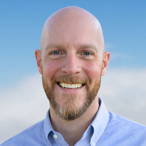 A smiling man with a beard and shaved head outdoors against a cloudy sky