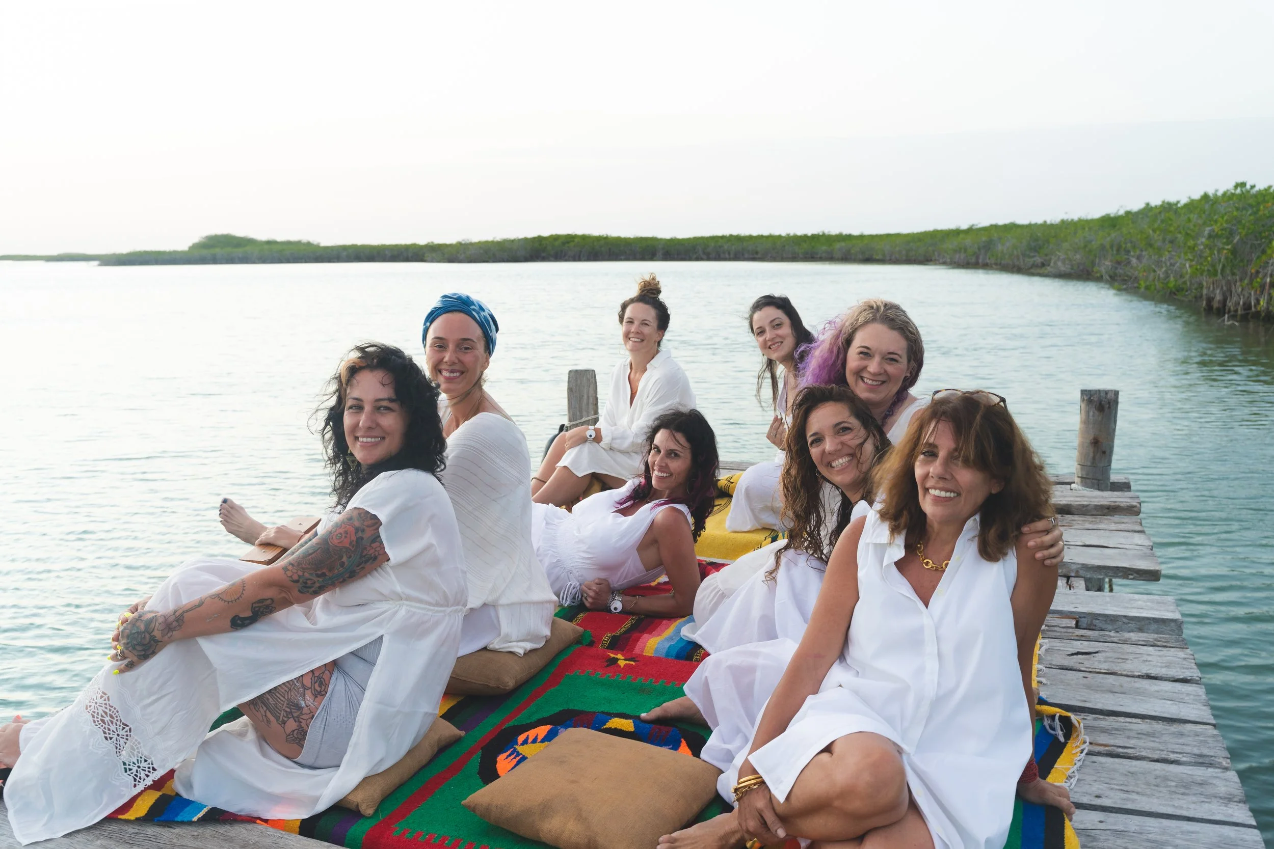 A group of women sitting on a colorful blanket on a wooden dock by the water, smiling at the camera. The background features calm water and a distant shoreline with greenery.