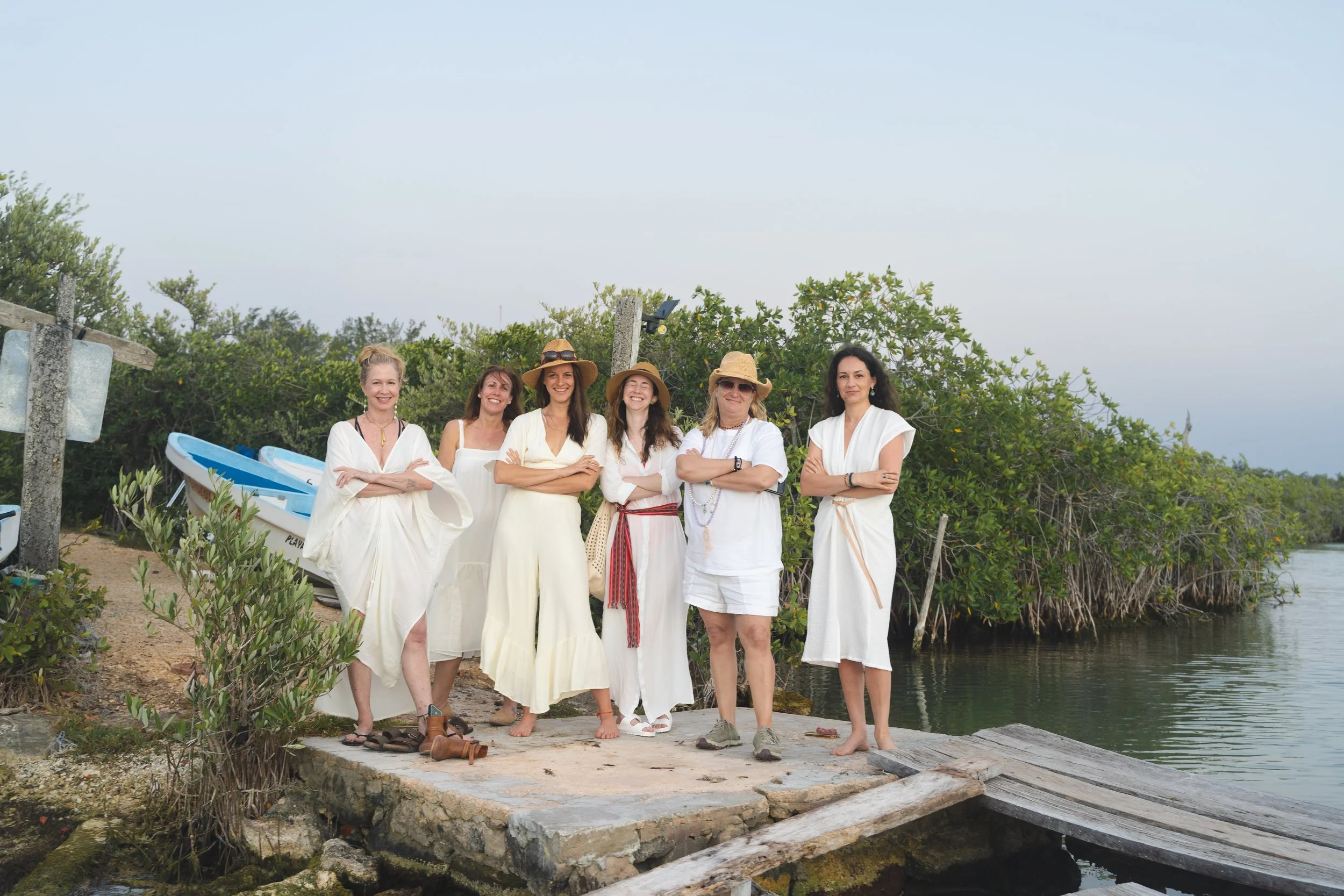 Group of six women standing on a dock near water, dressed in white, some wearing hats, with greenery and a boat in the background.