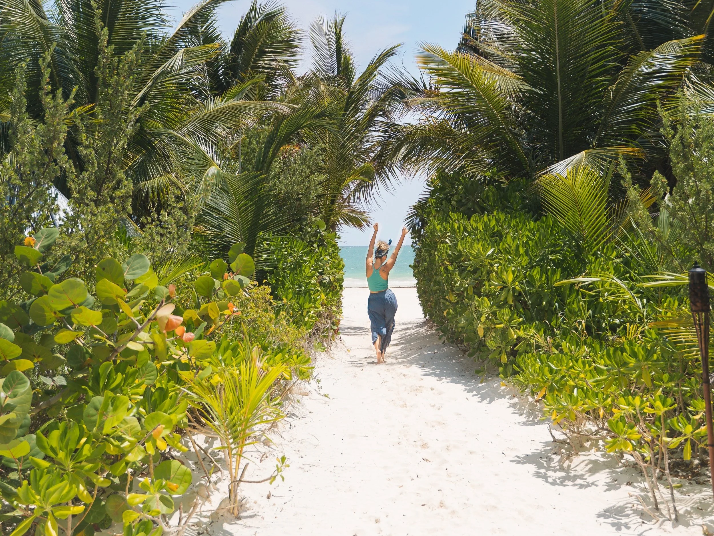A woman is walking through a sandy pathway bordered by lush green tropical plants and shrubs, with tall palm trees overhead and a view of the ocean in the distance.
