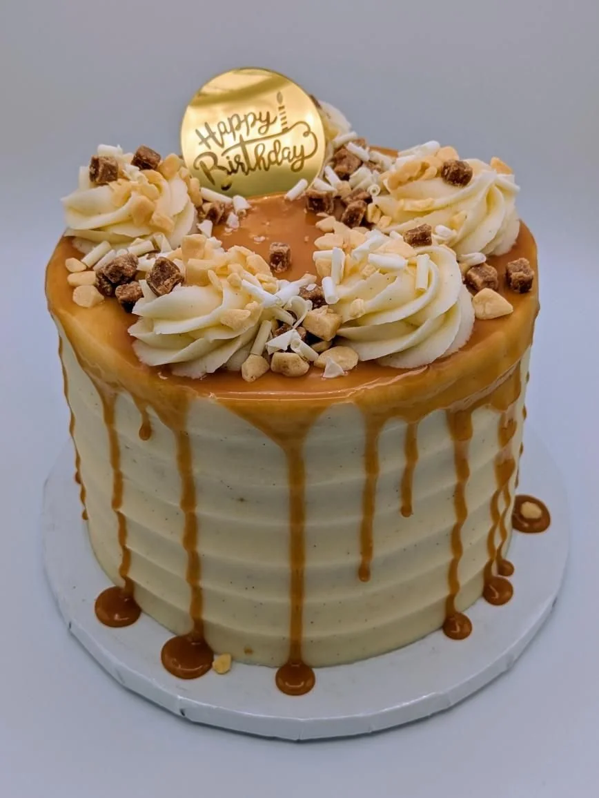 A birthday cake with white frosting, caramel drizzle on the sides, topped with white and chocolate sprinkles, and a gold "Happy Birthday" topper.