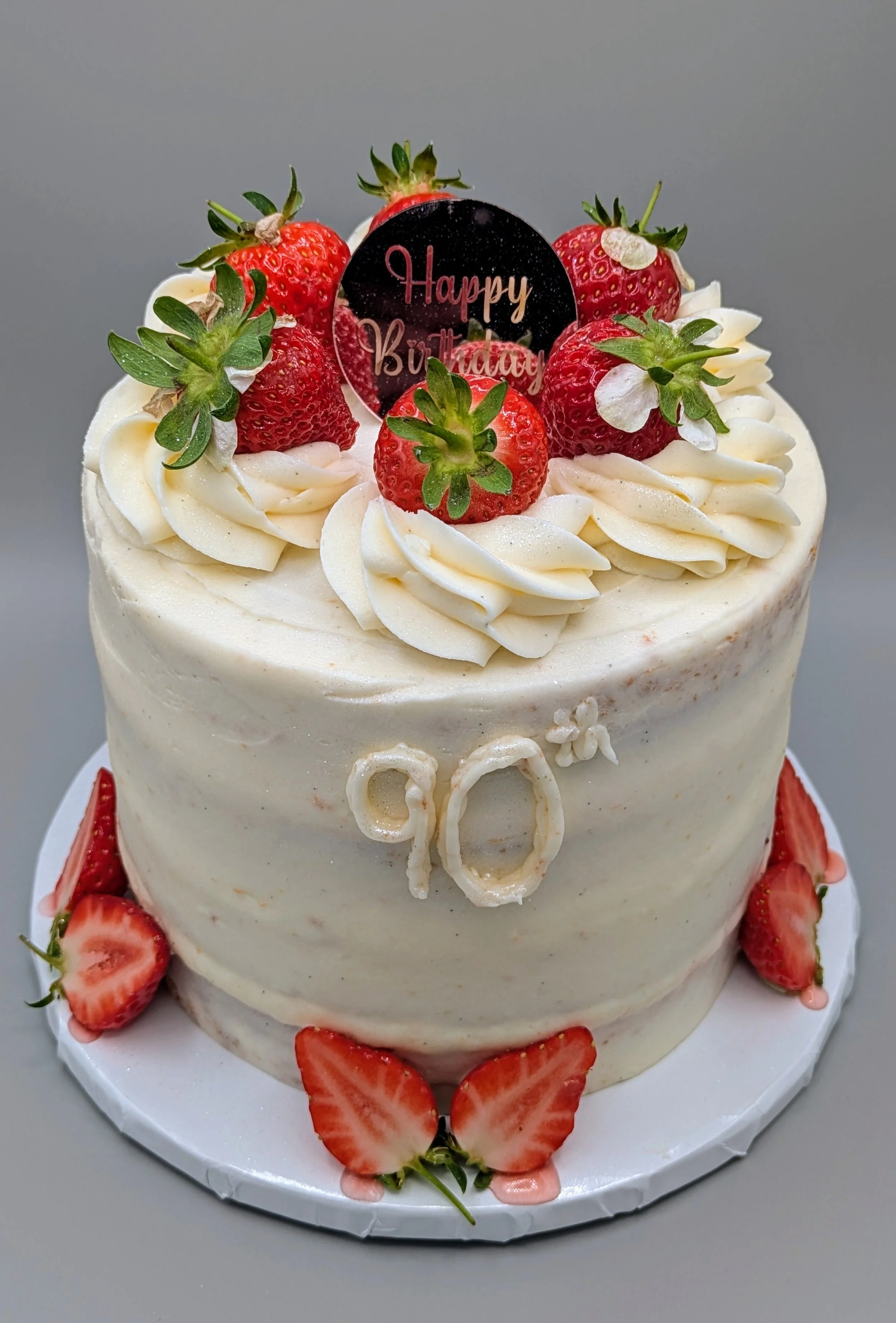 A white birthday cake decorated with strawberries and cream, with a "Happy Birthday" topper and the number 90 on the front.
