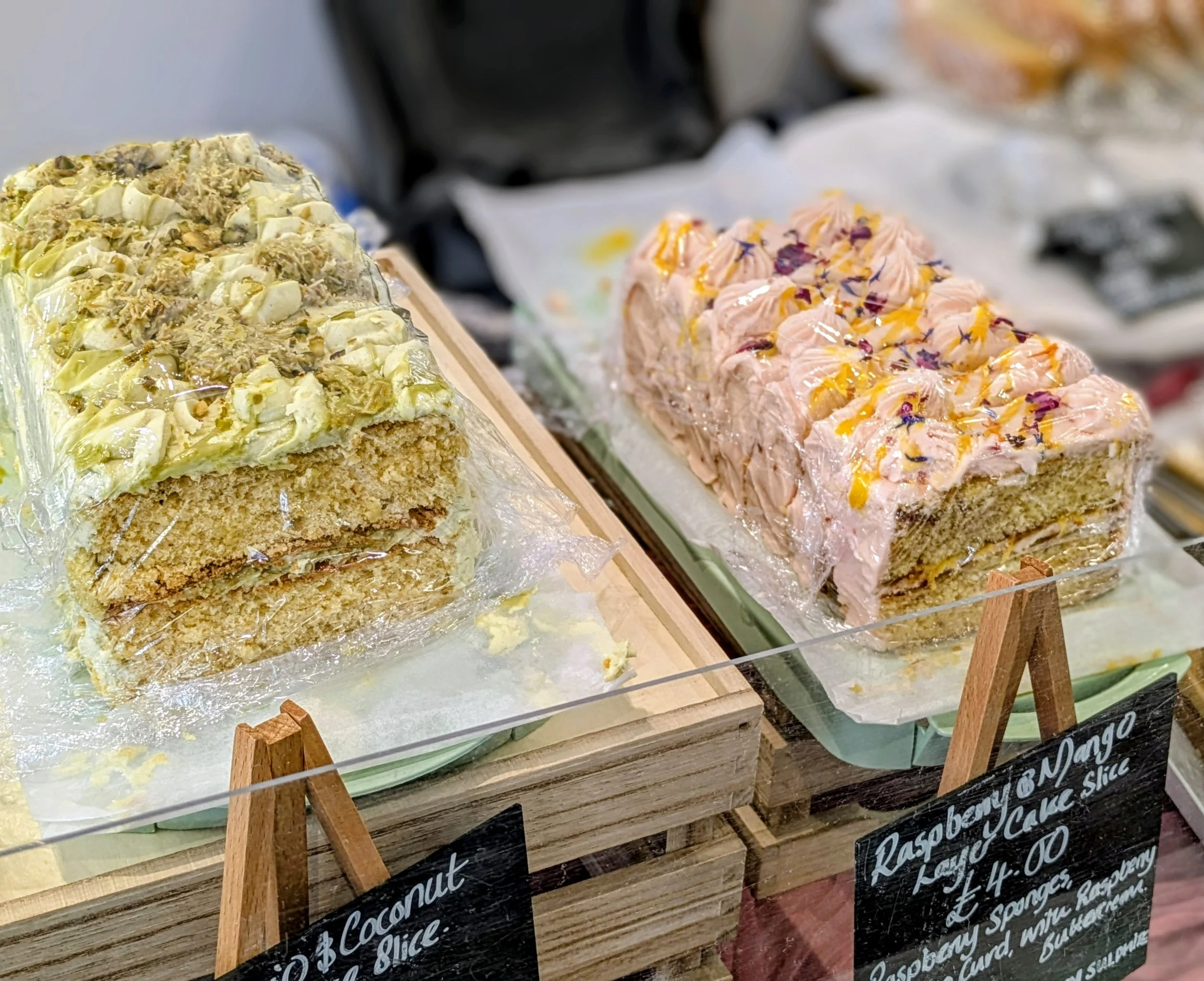 Two large slices of cake with frosting and toppings on display in a bakery, one coconut and the other raspberry and mango flavored.