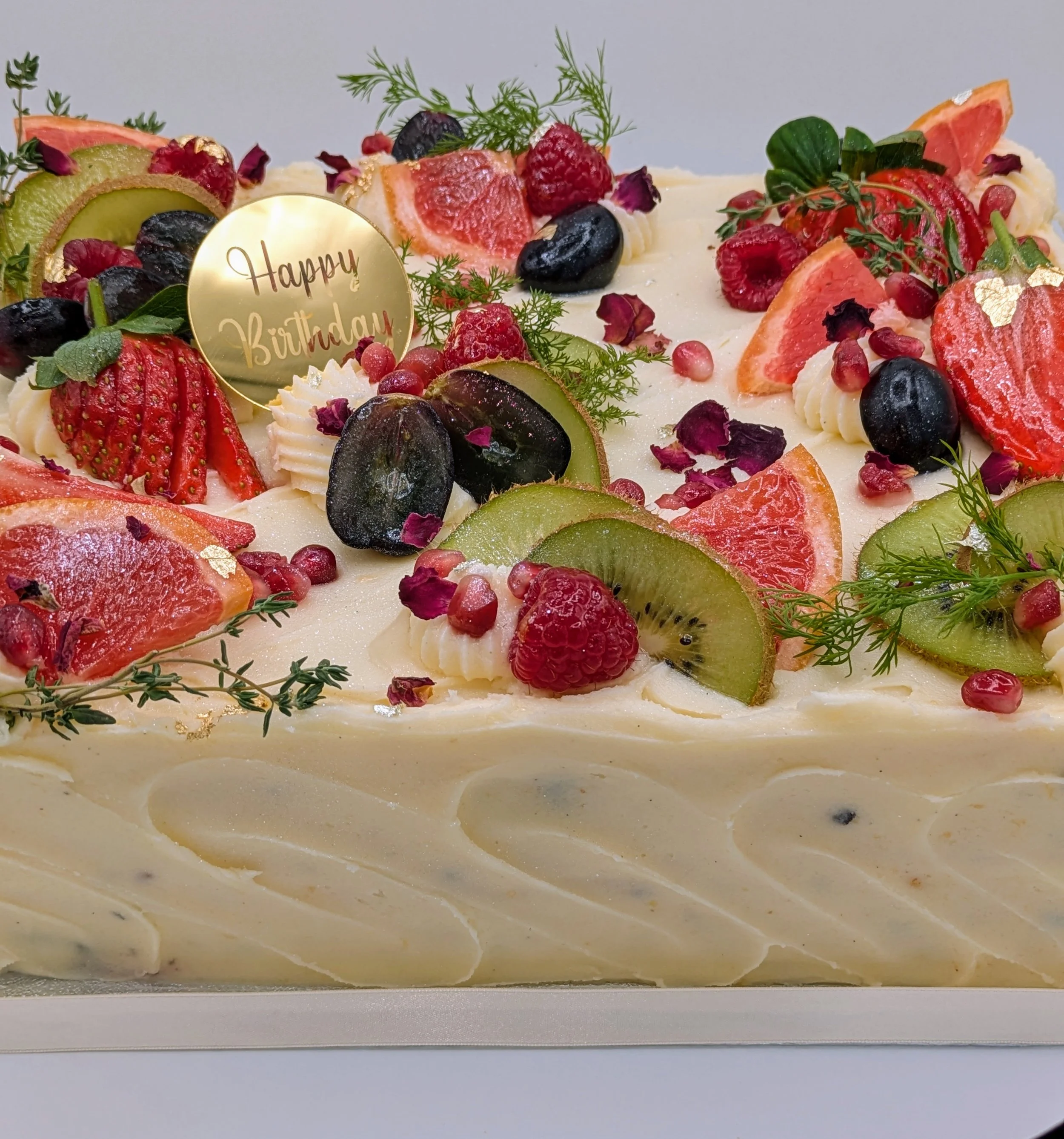 A decorated birthday cake with fresh fruit toppings including strawberries, kiwi, grapes, raspberries, and pomegranate seeds, with a gold "Happy Birthday" topper.