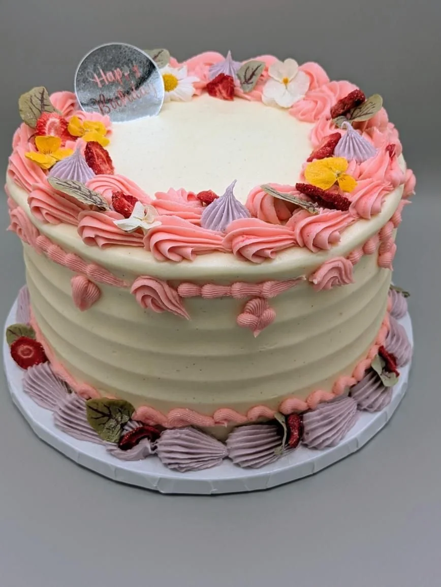 A round birthday cake with white frosting, decorated with pink swirls and small purple and yellow frosting flowers, topped with edible leaves, red berries, and a message reading 'Happy Birthday' on a clear topper.