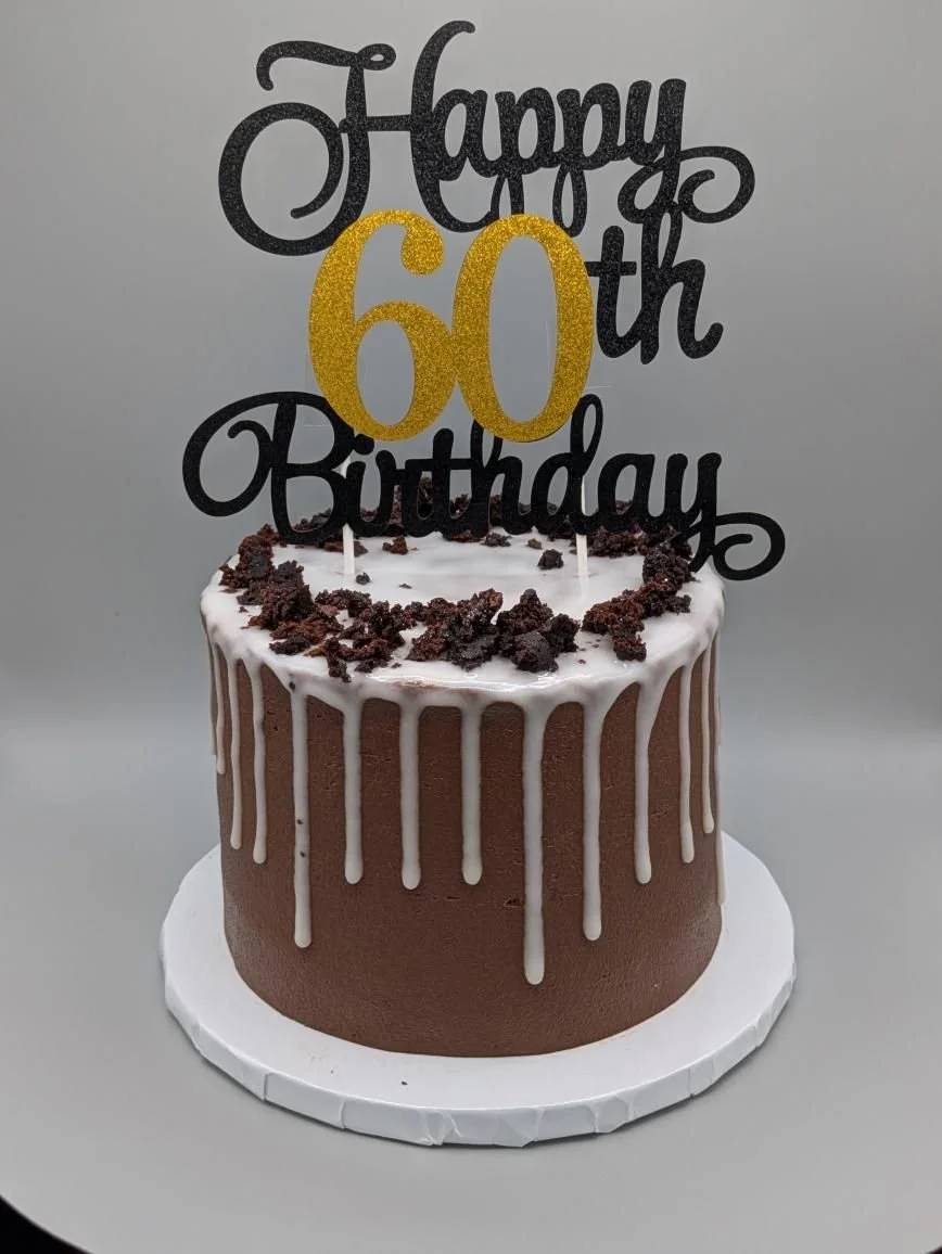 A chocolate birthday cake with white drip icing, topped with crumbled cookies and a black and gold 'Happy 60th Birthday' cake topper.