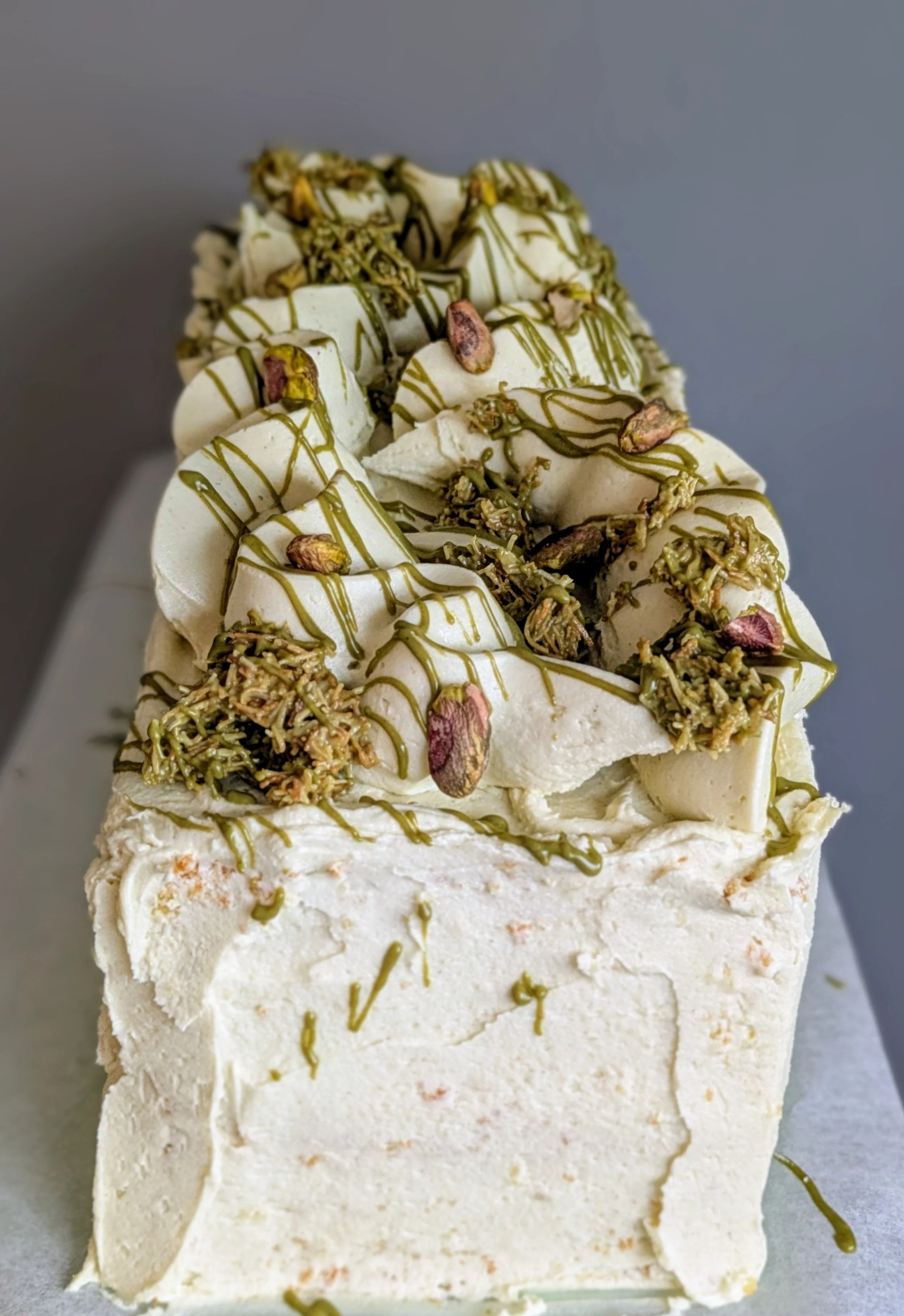 A rectangular slice of white ice cream cake topped with chopped pistachios, rose petals, and drizzled with green sauce.