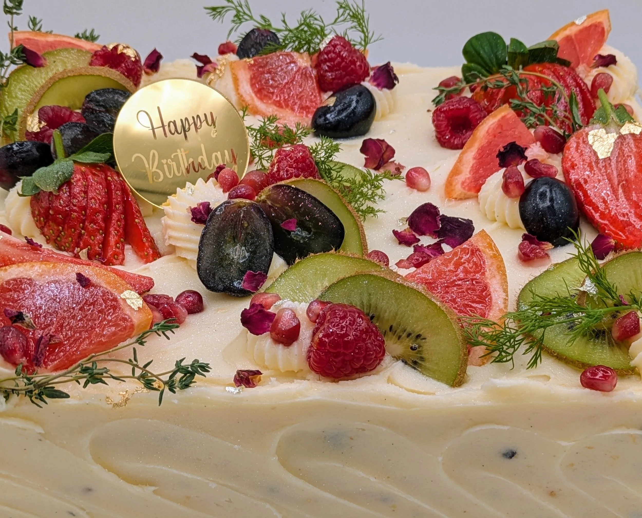 A birthday cake decorated with various fresh fruits including strawberries, blueberries, kiwi, grapefruit, and pomegranate seeds, with sprigs of herbs and a gold circle plaque that says 'Happy Birthday'.