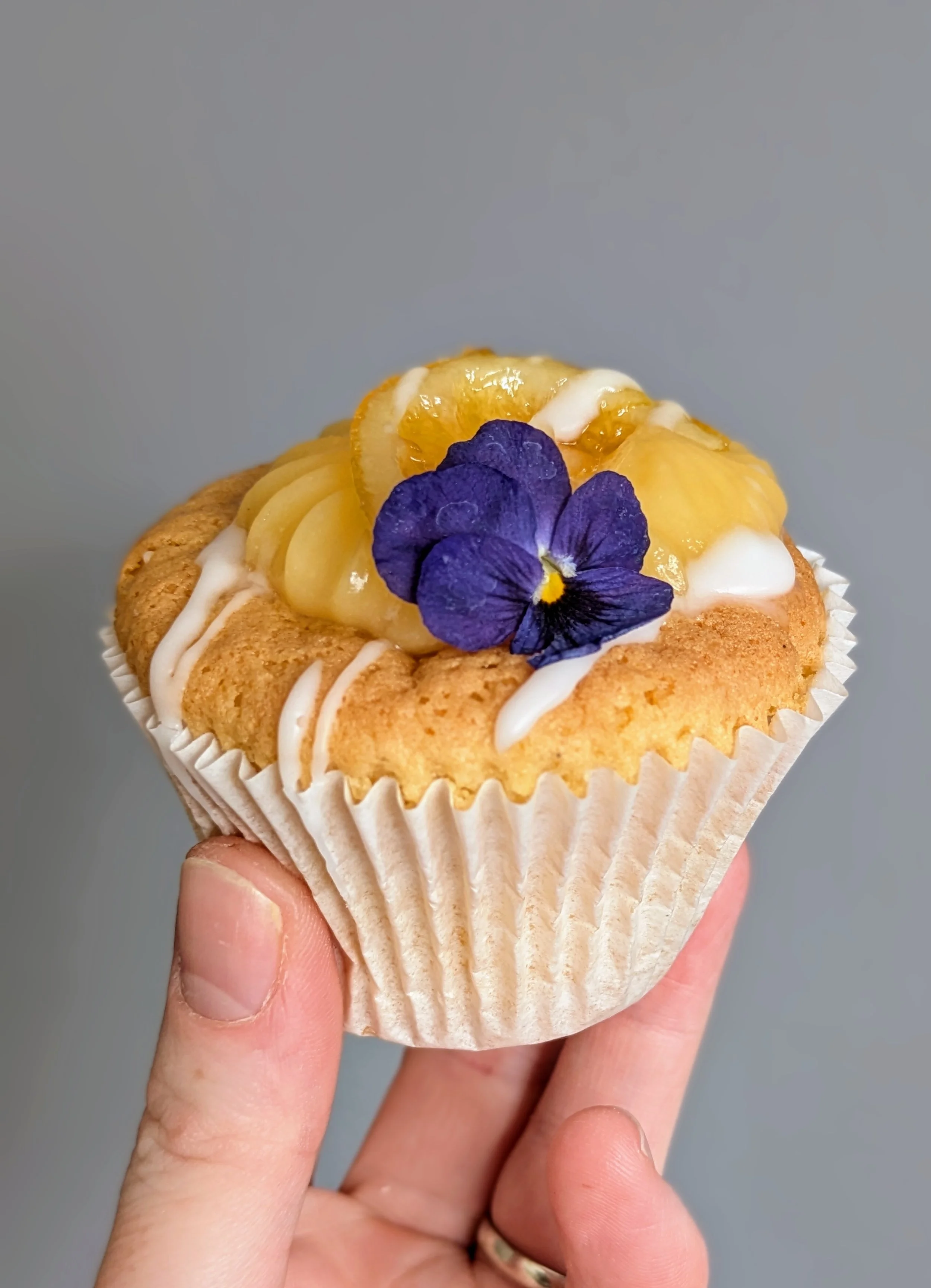 Hand holding a cupcake topped with lemon slices, a purple edible flower, and white icing drizzle, with a plain gray background.