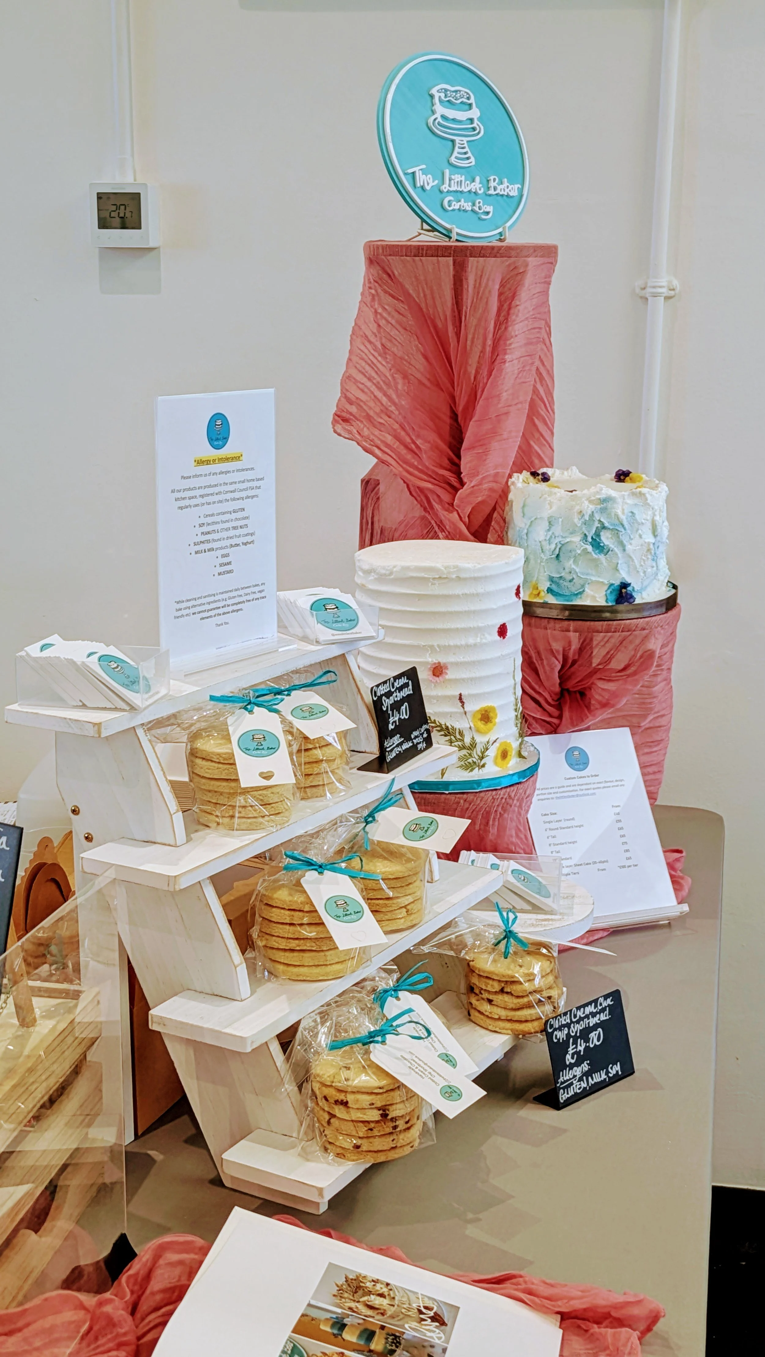 Display of baked goods at a bakery counter, including jars of cookies, decorated cakes, and a sign for The Littlest Baker, with a pink fabric backdrop and a menu on a stand.