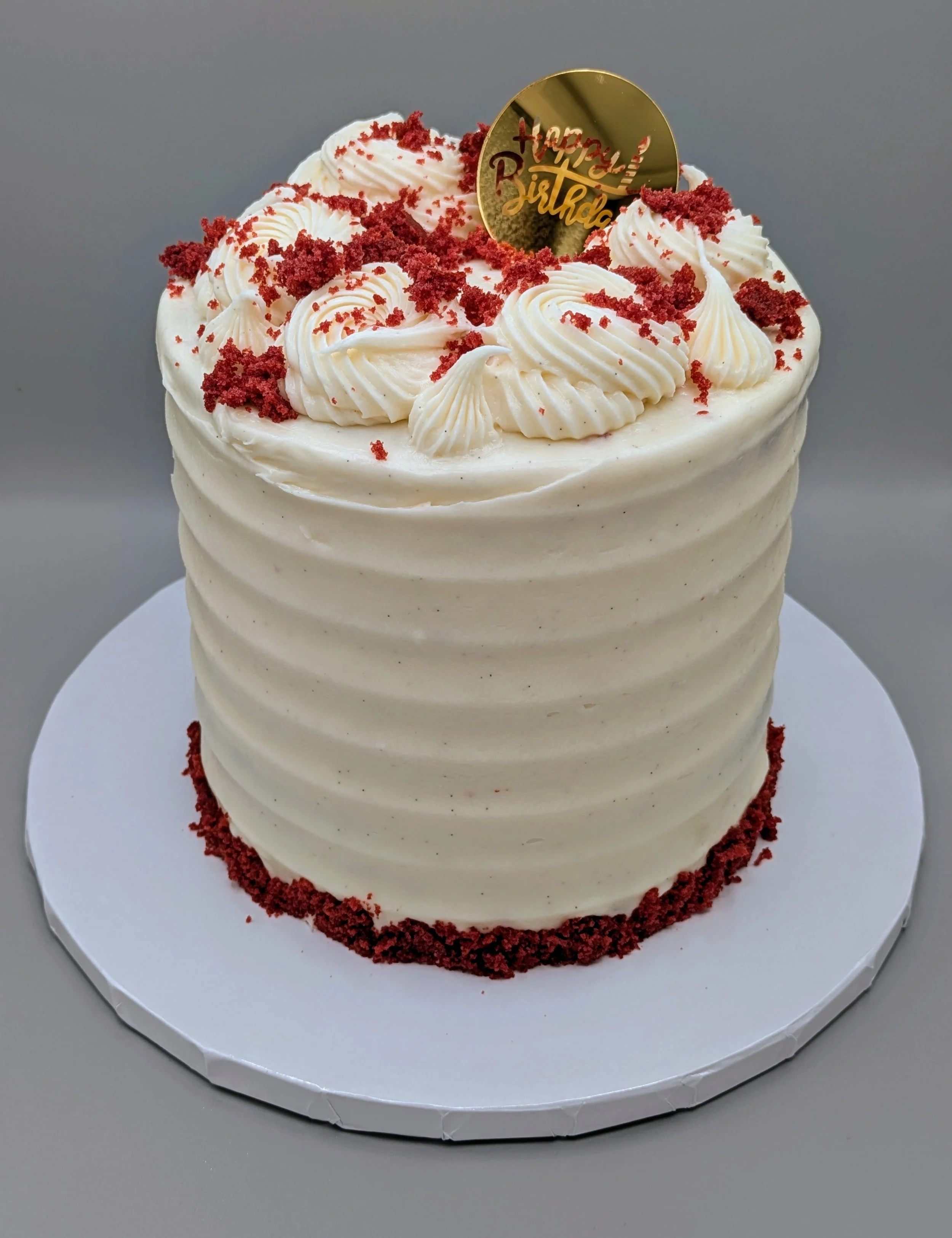 A tall white birthday cake decorated with red crumbs and swirls of white frosting on top, with a gold 'Happy Birthday' topper.