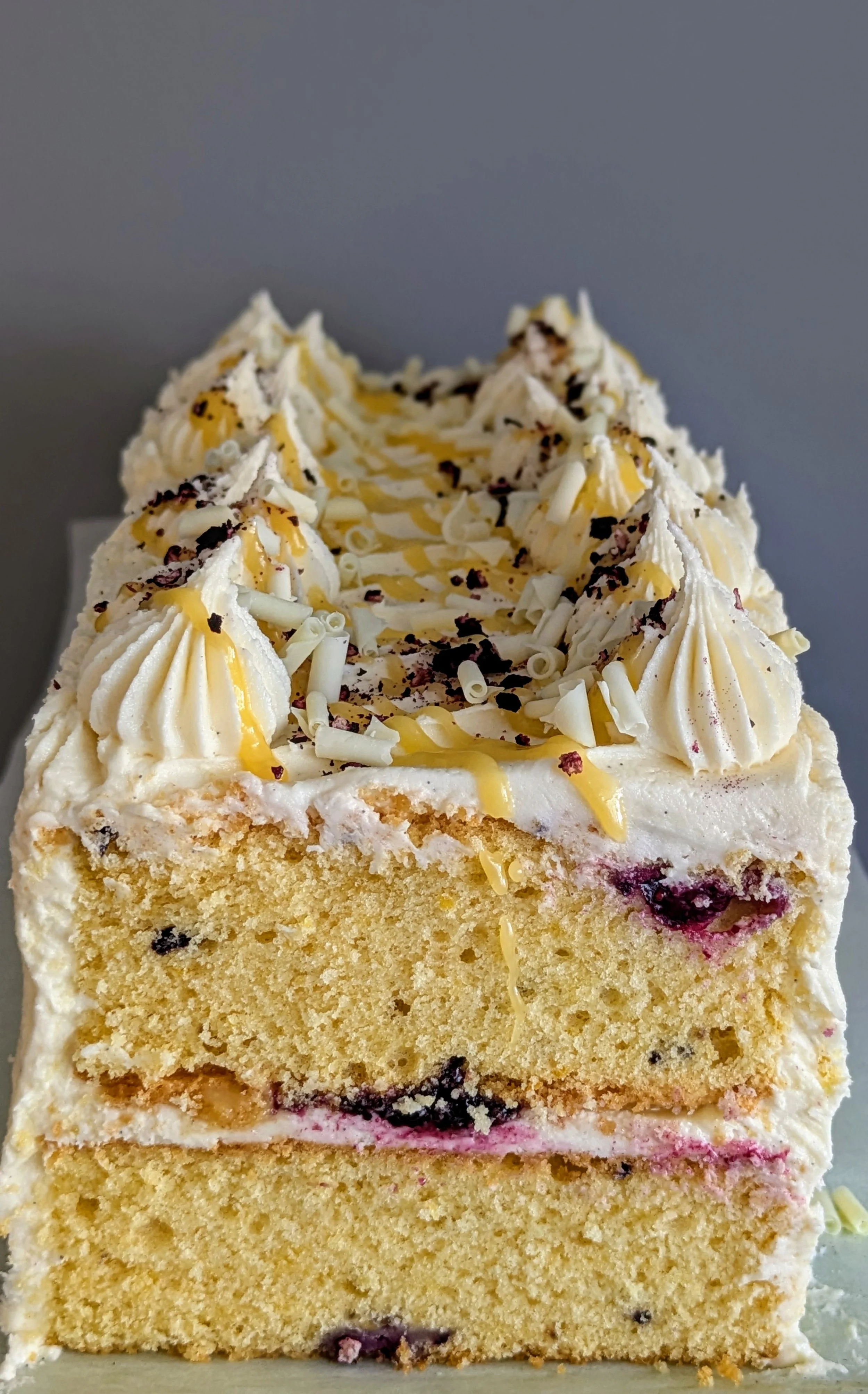 Close-up of a layered vanilla cake with white frosting, decorated with white chocolate curls, chocolate shavings, and caramel drizzle on top.