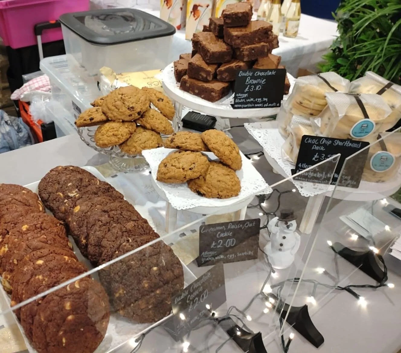 Assorted cookies and brownies on display at a bakery or market stall, including chocolate chip cookies, flapjack bars, and double chocolate brownies, with handwritten price signs.