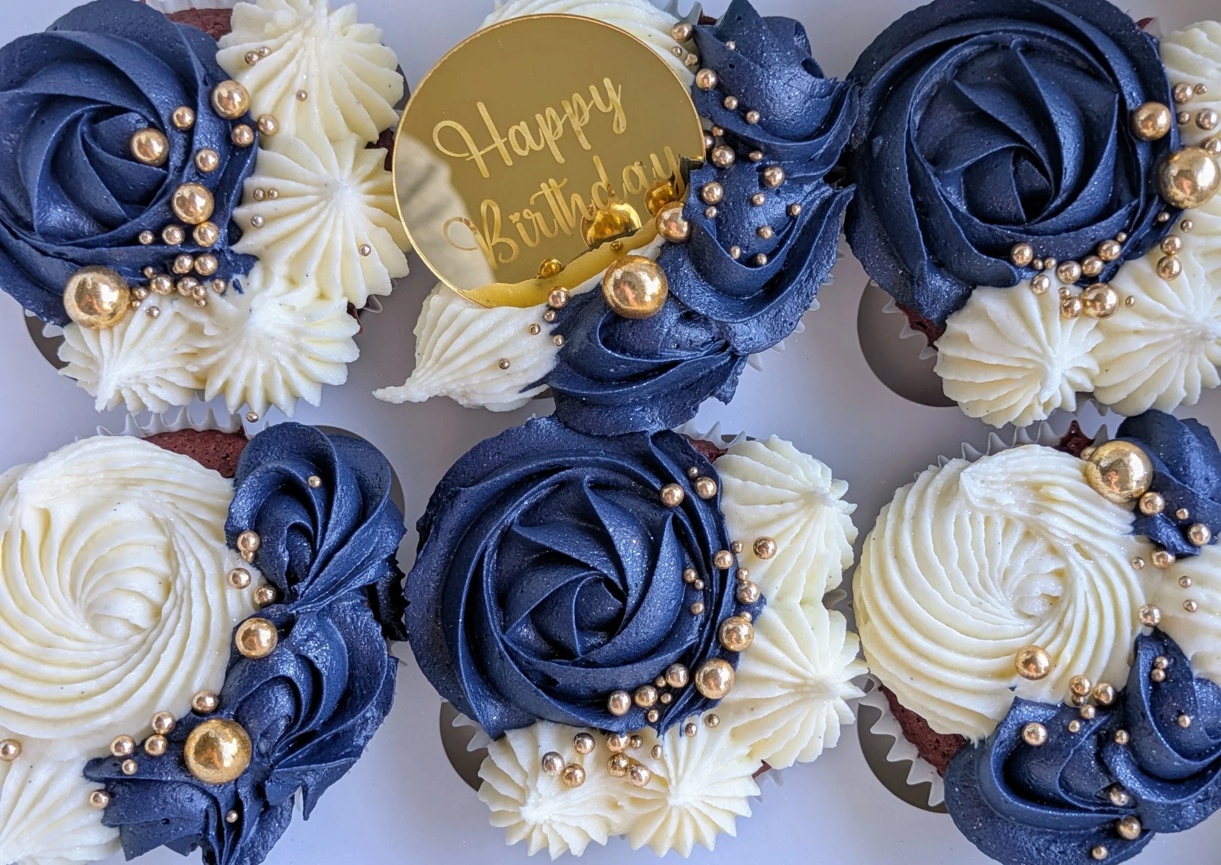 Cupcakes decorated with blue and white swirled frosting, adorned with gold-colored edible beads, and a gold 'Happy Birthday' topper.