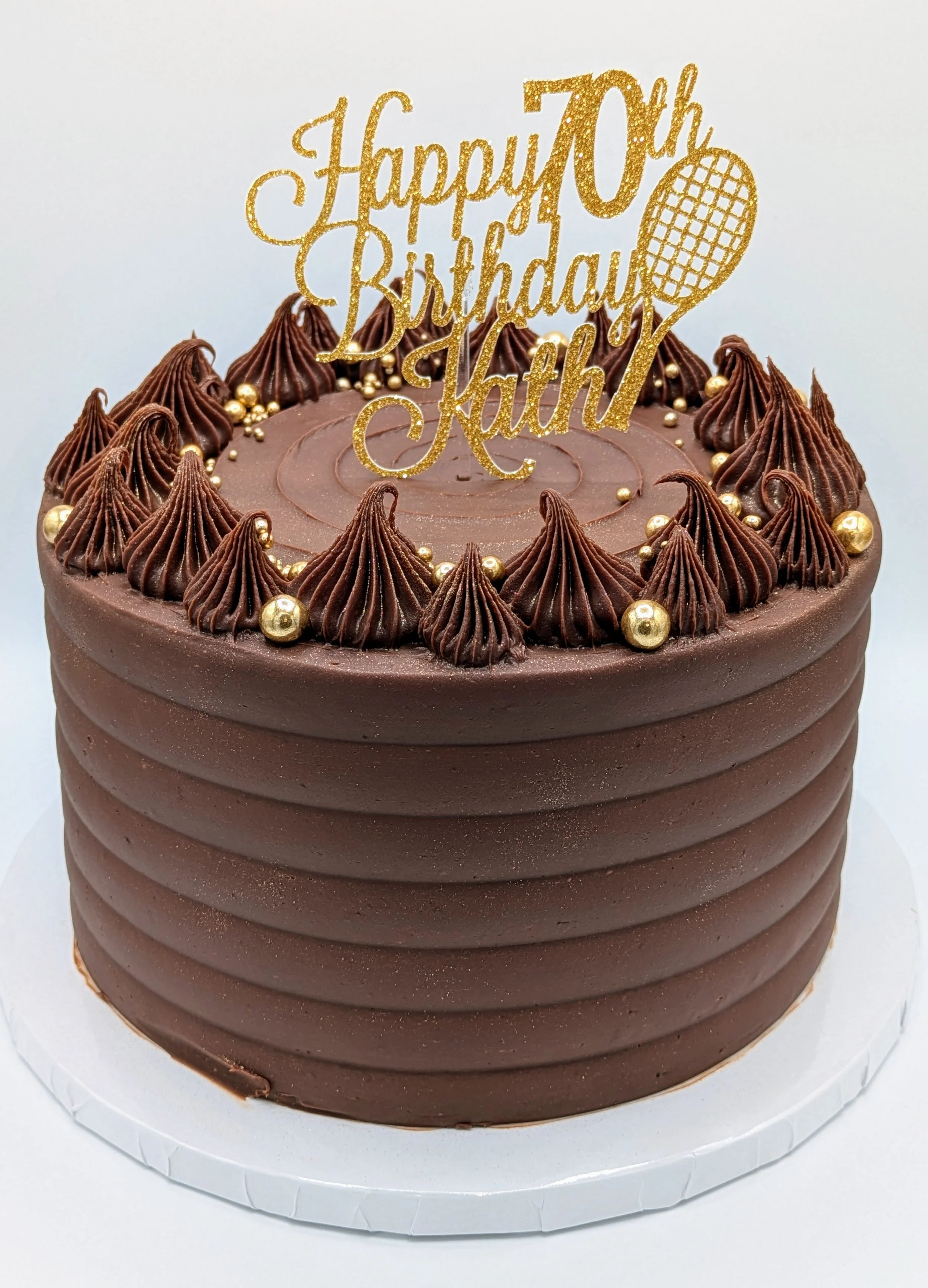 Chocolate birthday cake with brown frosting, decorated with rosettes and gold pearls, and a gold 'Happy 70th Birthday Kath' topper with a tennis racket illustration.