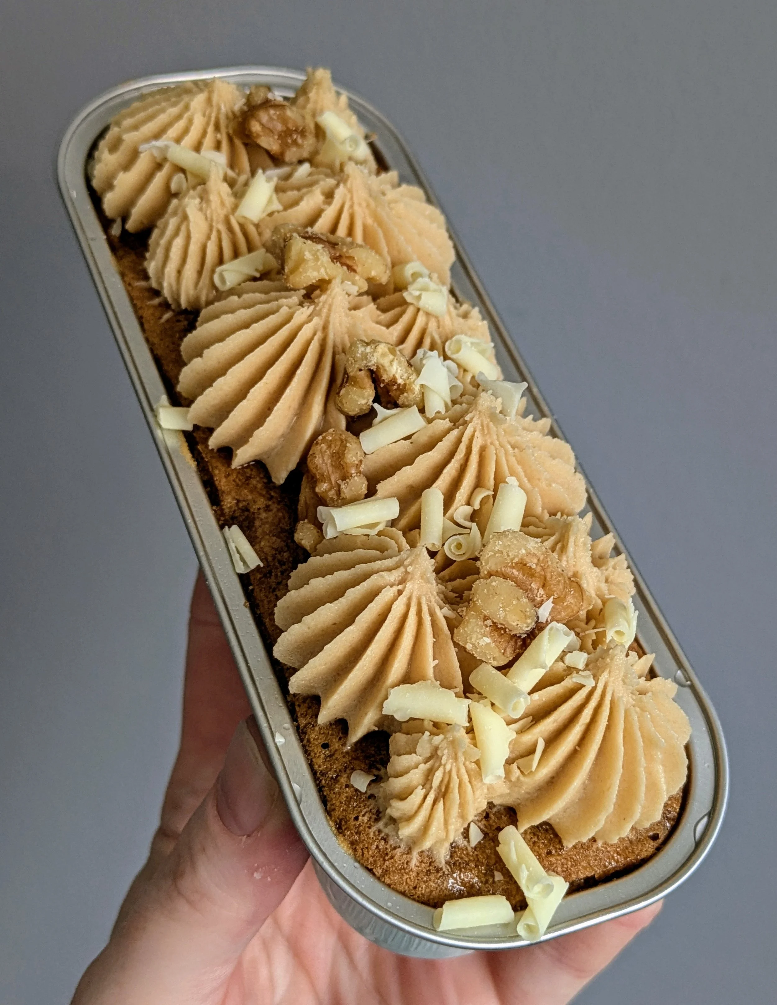 A hand holding a rectangular dessert with piped caramel frosting, white chocolate shavings, and caramelized nuts on top.