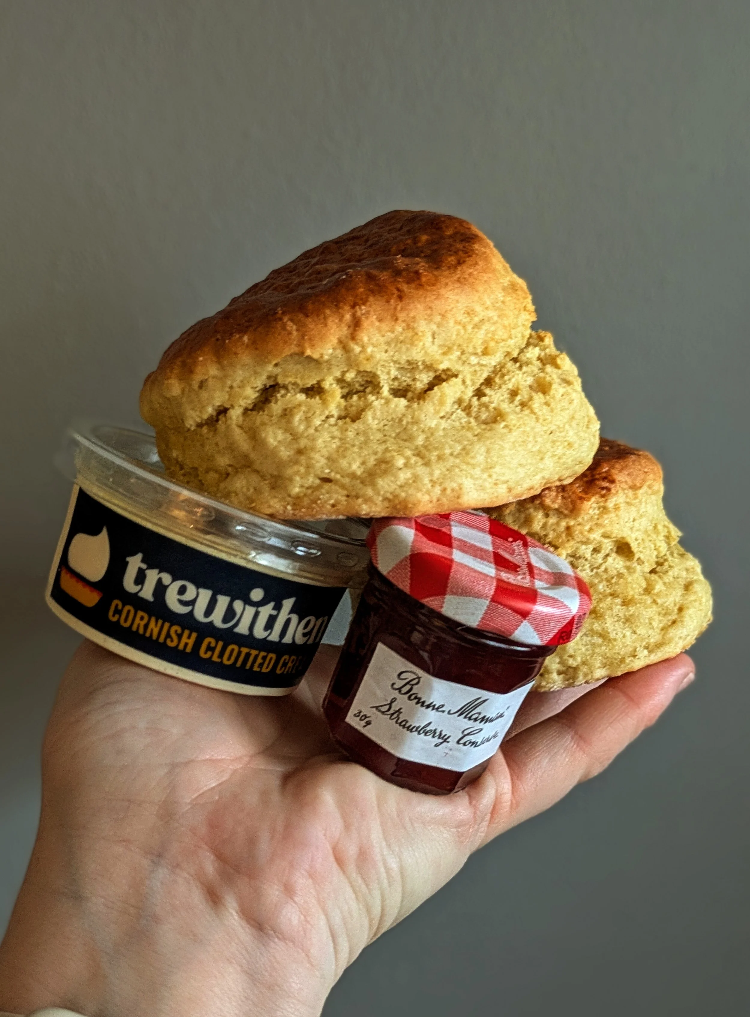 Hand holding two scones, a jar of strawberry jam, and a plastic container of Cornish clotted cream from Trewithen against a plain background.