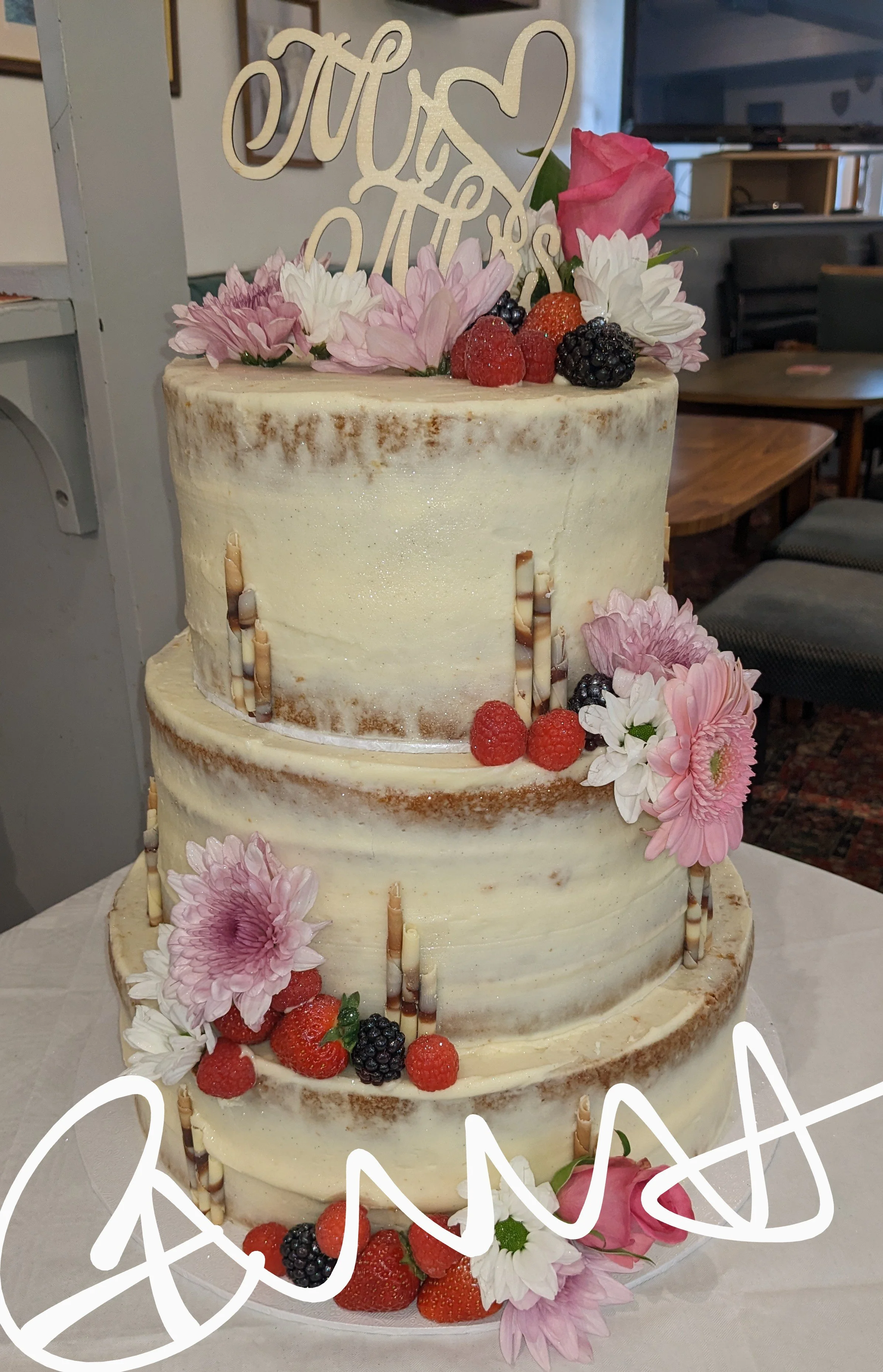 Three-tiered wedding cake decorated with pink and white flowers, fresh berries, and vertical chocolate accents, with a tiered topper that says 'Love' with a heart.