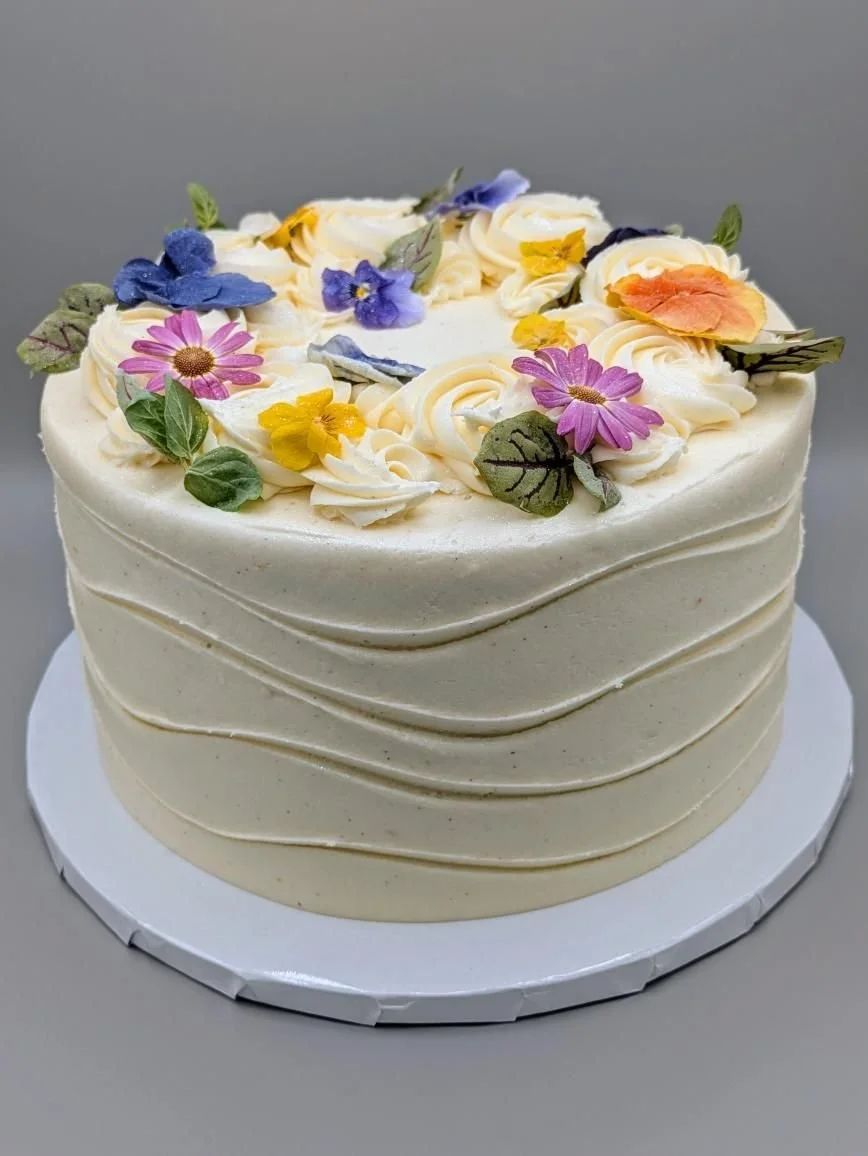 Cream-colored cake with wave frosting and colorful edible flower decorations on top, placed on a white cake board.