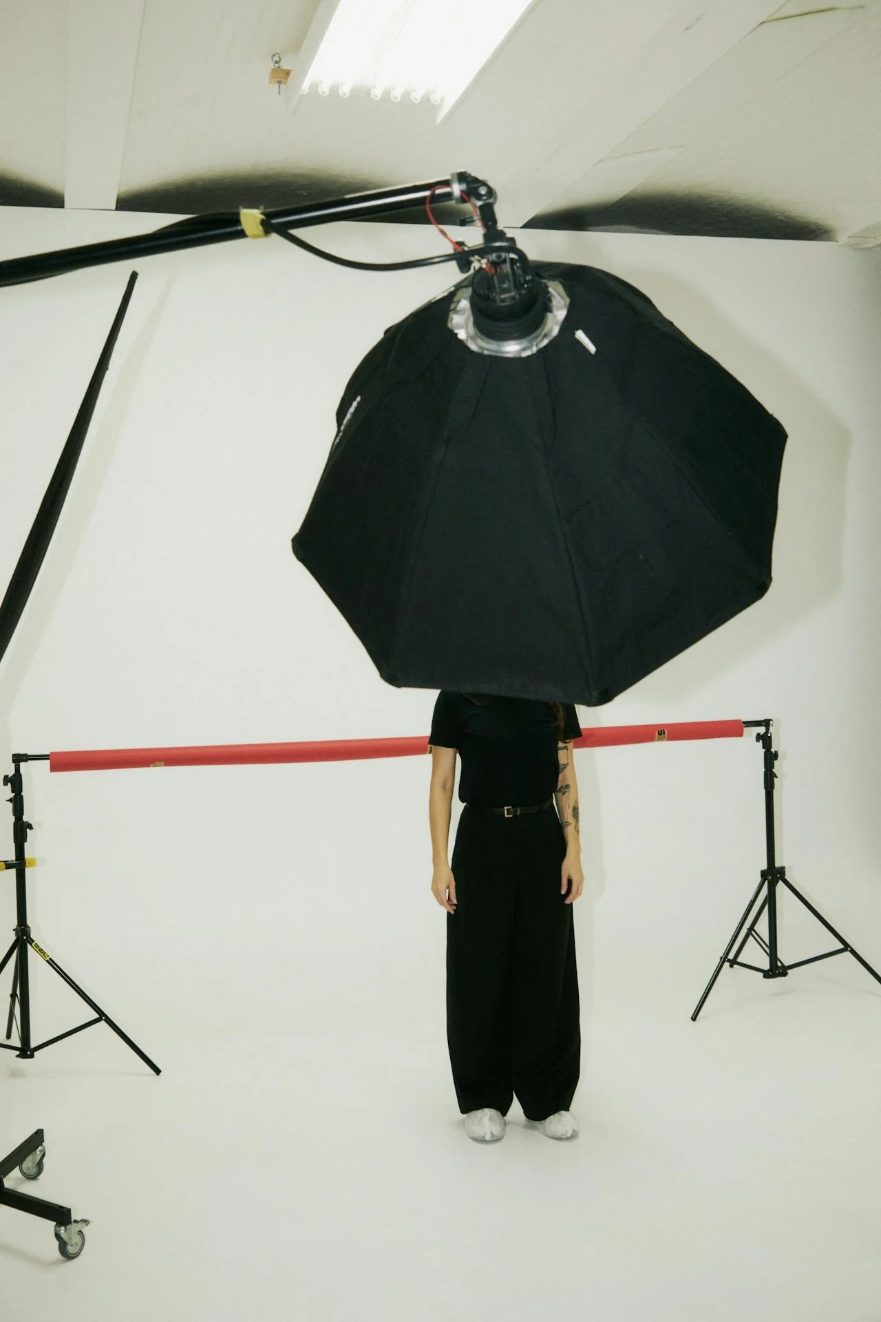 Person partially obscured by large black umbrella light in a photography studio, with red backdrop bar in background.