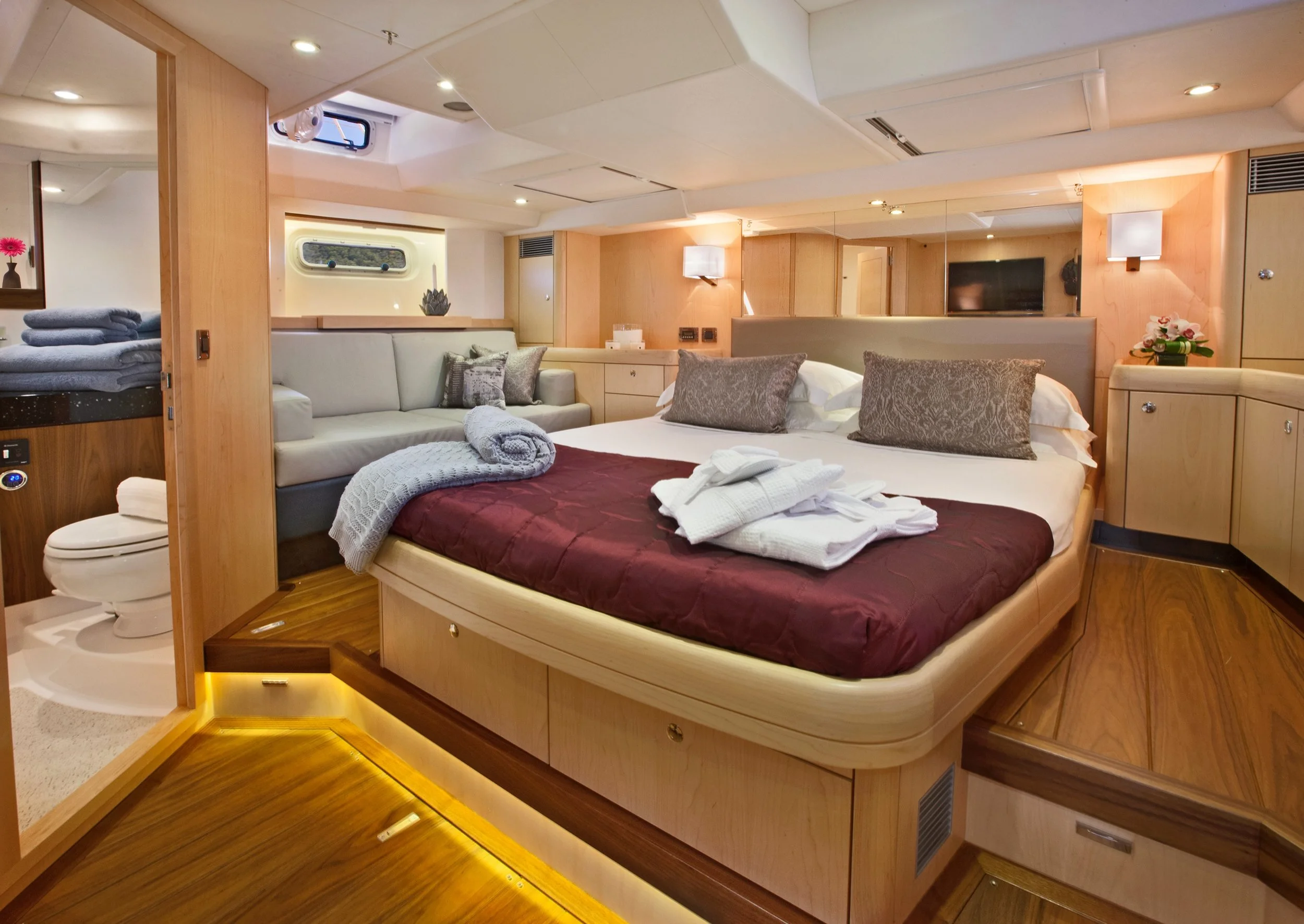 Luxury bedroom with a large bed, decorative pillows, and a folded robe on top. A sitting area with a sofa and towels, a small bathroom with a toilet, and wooden and modern decor with soft lighting.