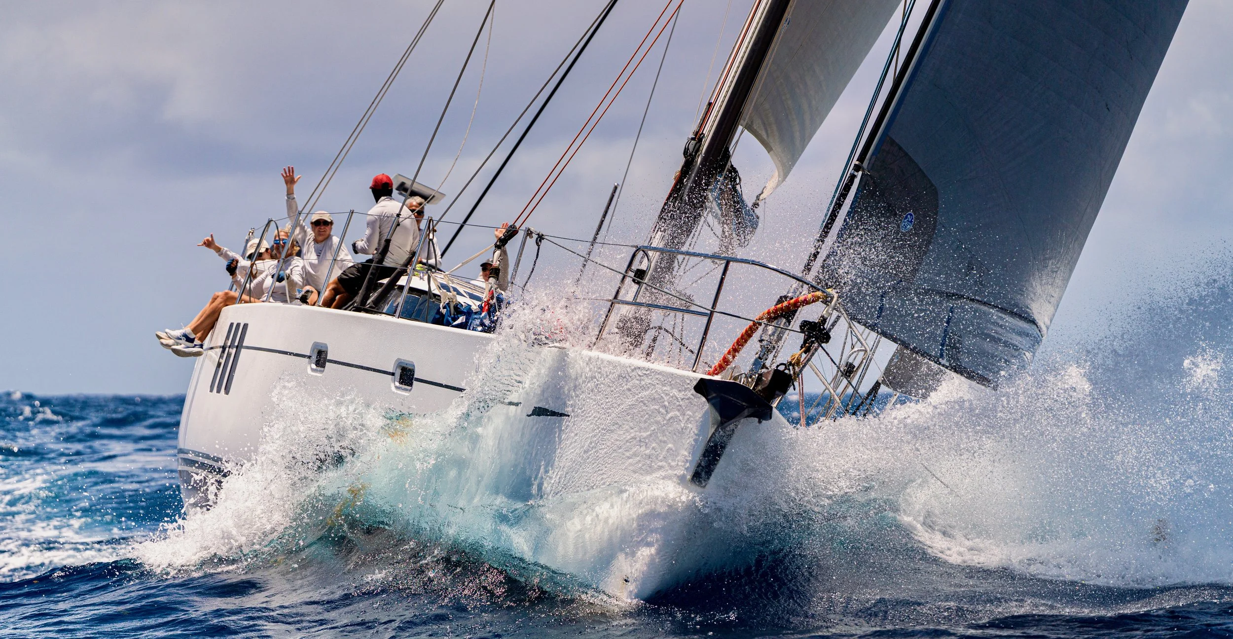 A sailboat speeding through the ocean with several people on board, some waving, as it cuts through the water creating a splash and foam.