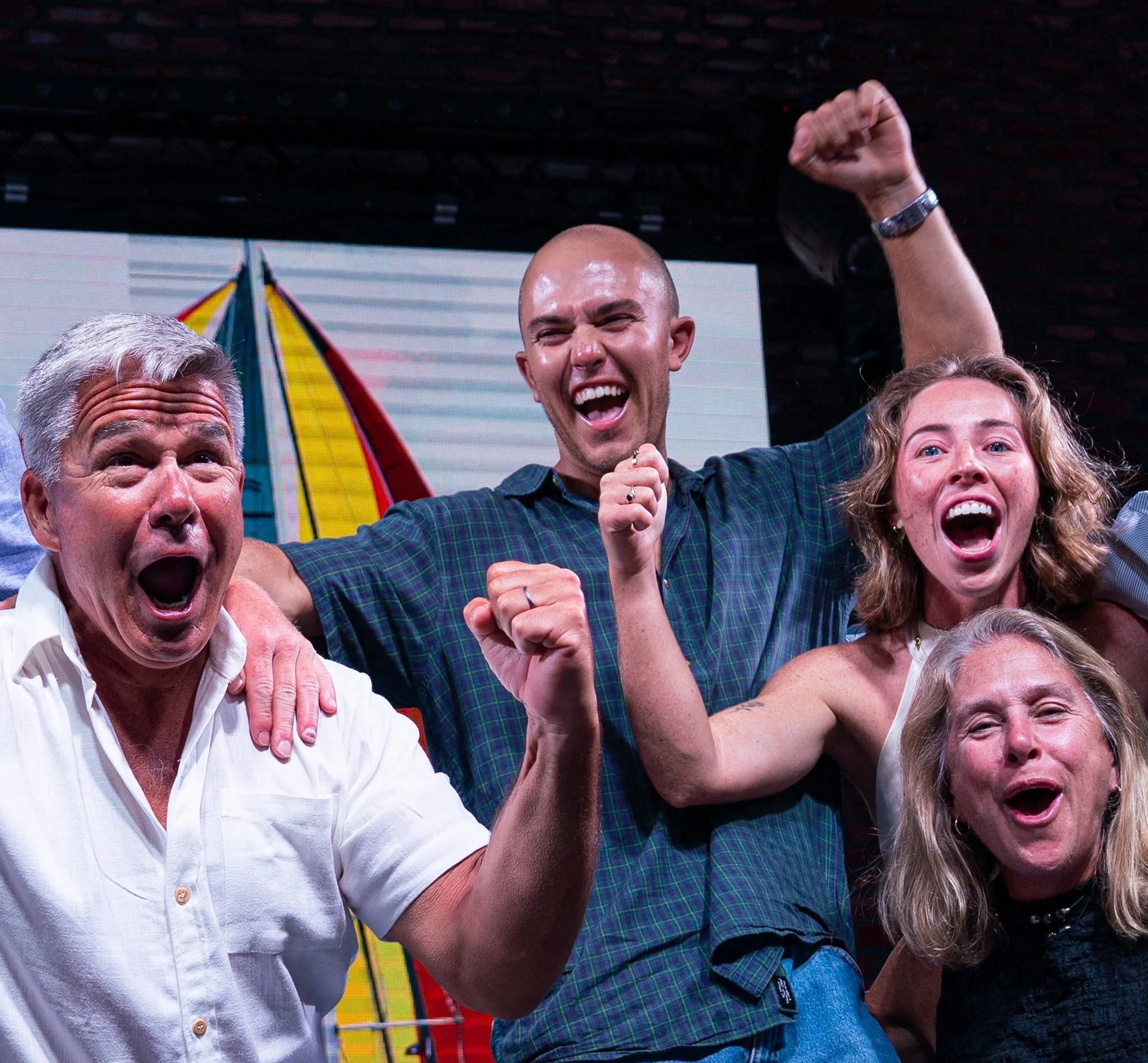 Group of people celebrating joyfully with raised fists, smiling faces, and open mouths, with a surfboard in the background.