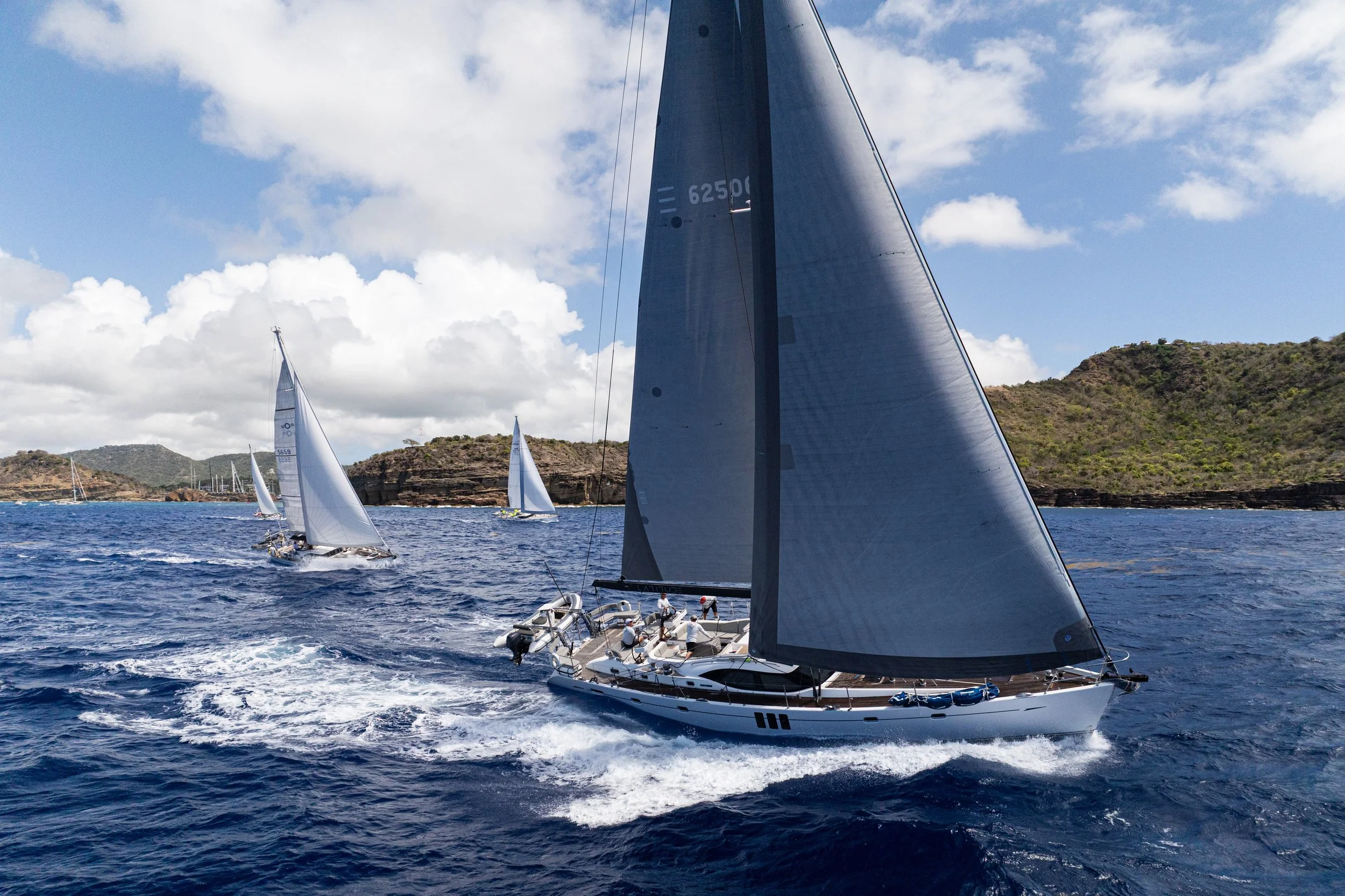 Sailboats racing on the ocean near a coastline with green hills and partly cloudy sky.