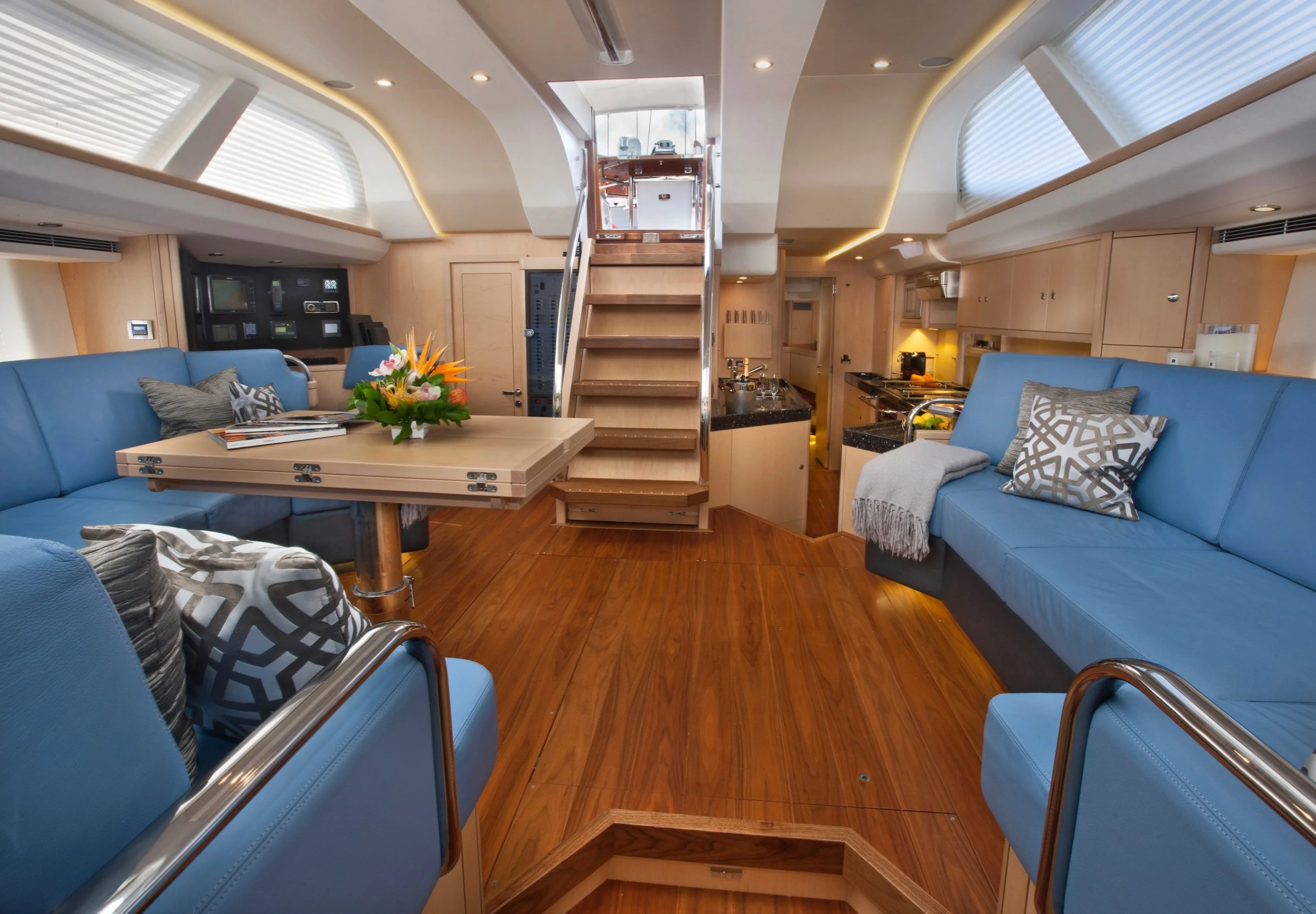 Interior of a yacht with blue sofas, a wooden floor, a central dining table with a flower arrangement, and a kitchen area in the background. A staircase leads to the upper deck.