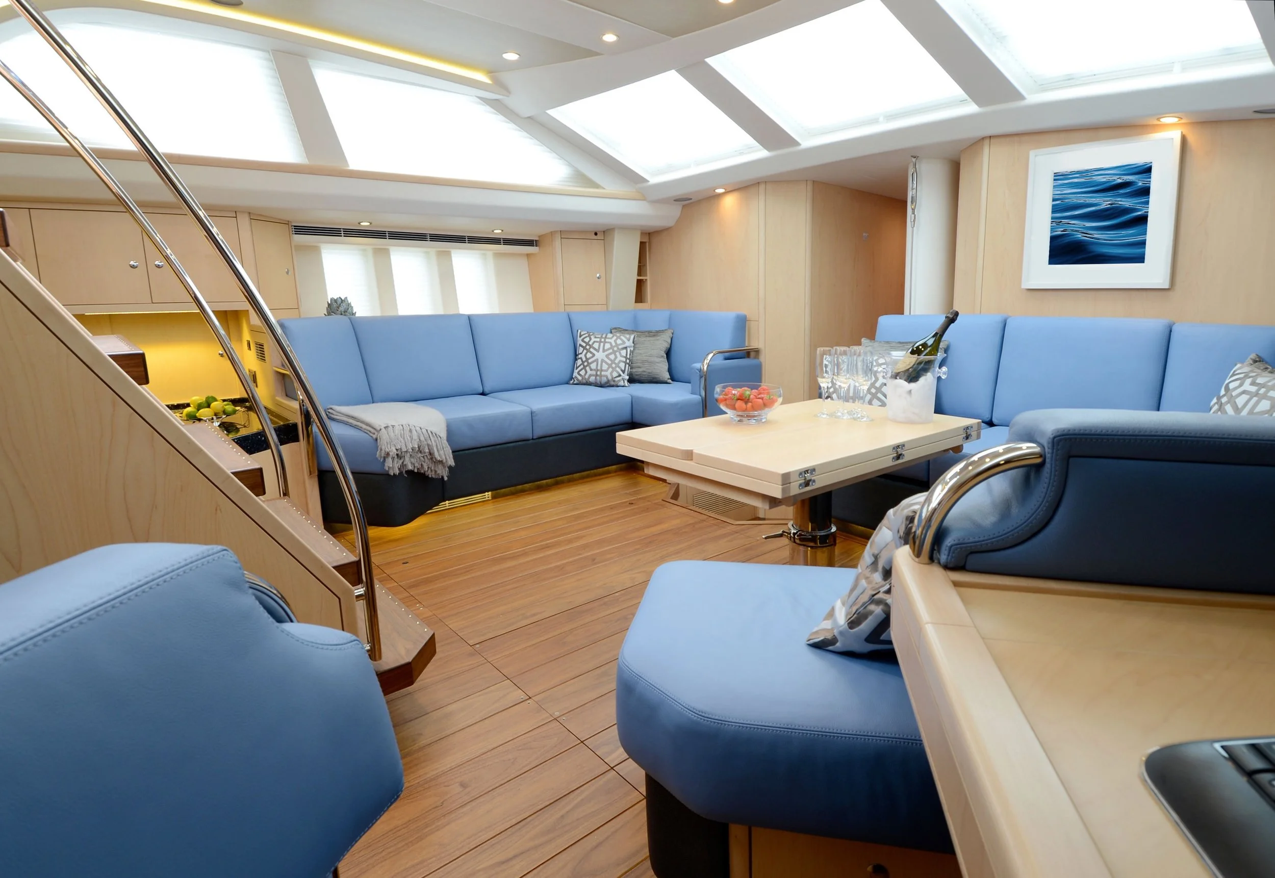 Luxurious yacht interior with blue seating, wooden floors, skylight ceiling, and a framed ocean picture on the wall.