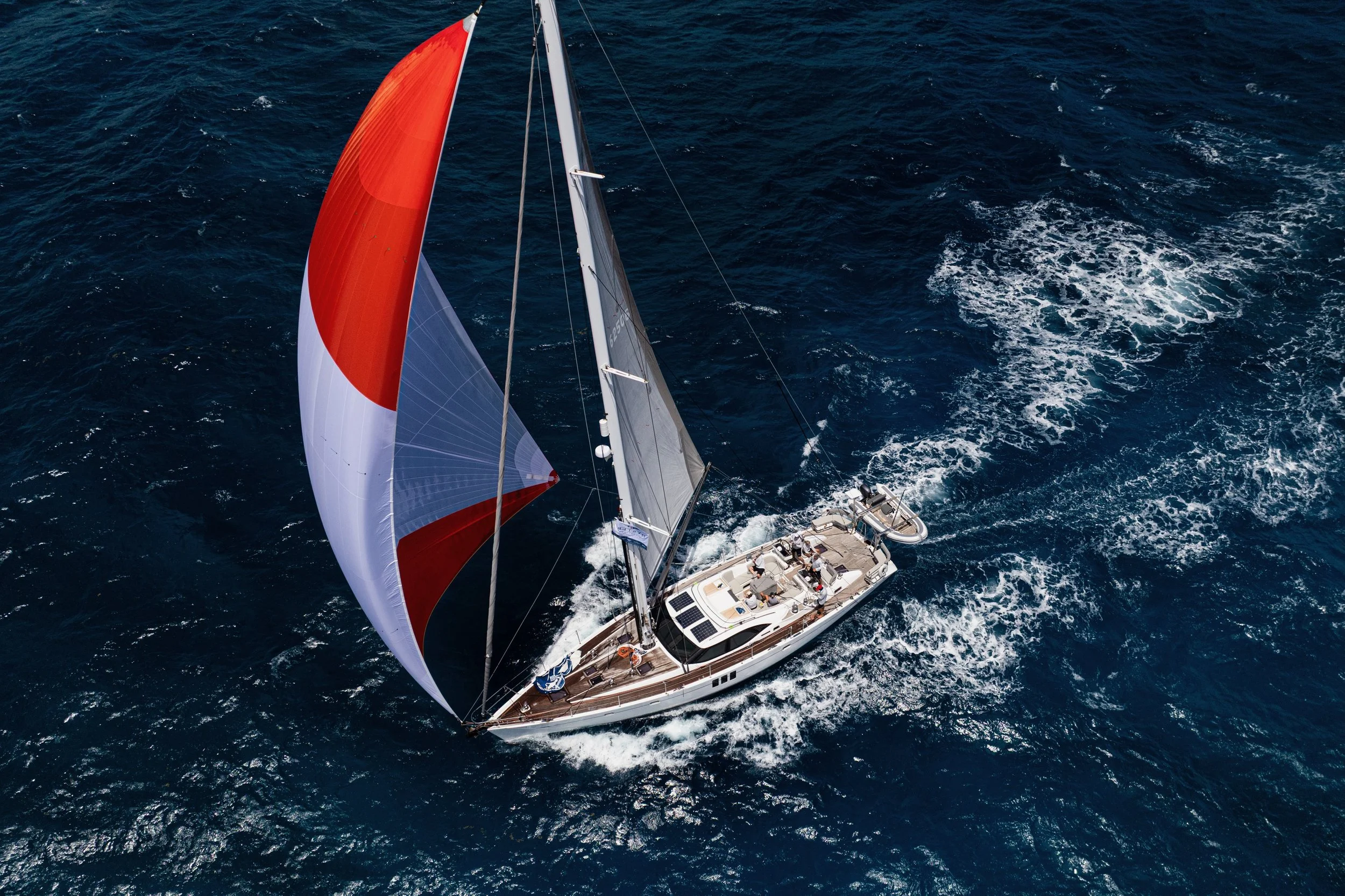 Overhead view of a sailing yacht with a red, white, and gray spinnaker sail, navigating through open ocean waters, leaving a wake behind.