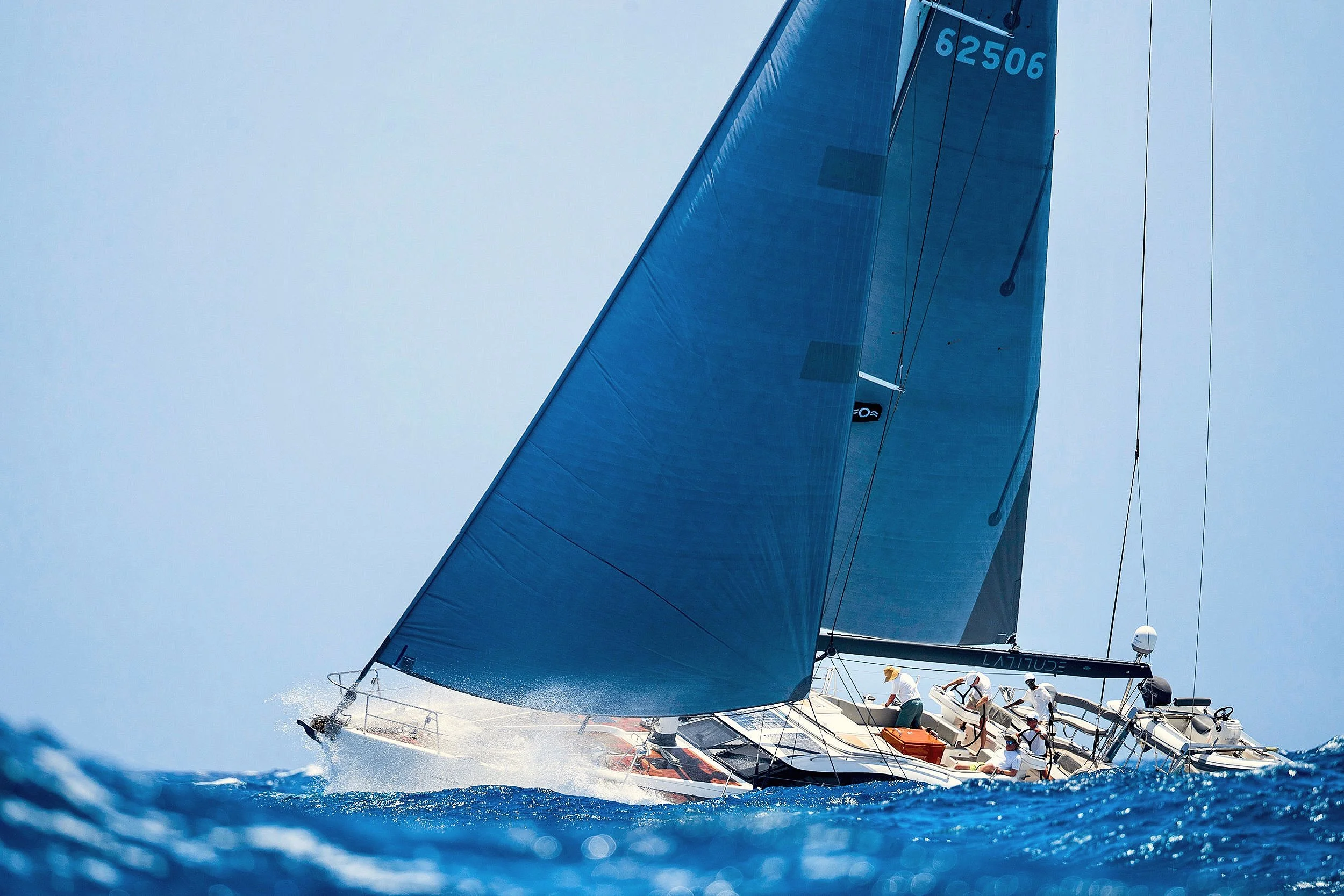 A sailboat with blue sails cutting through blue ocean water with crew members on board.