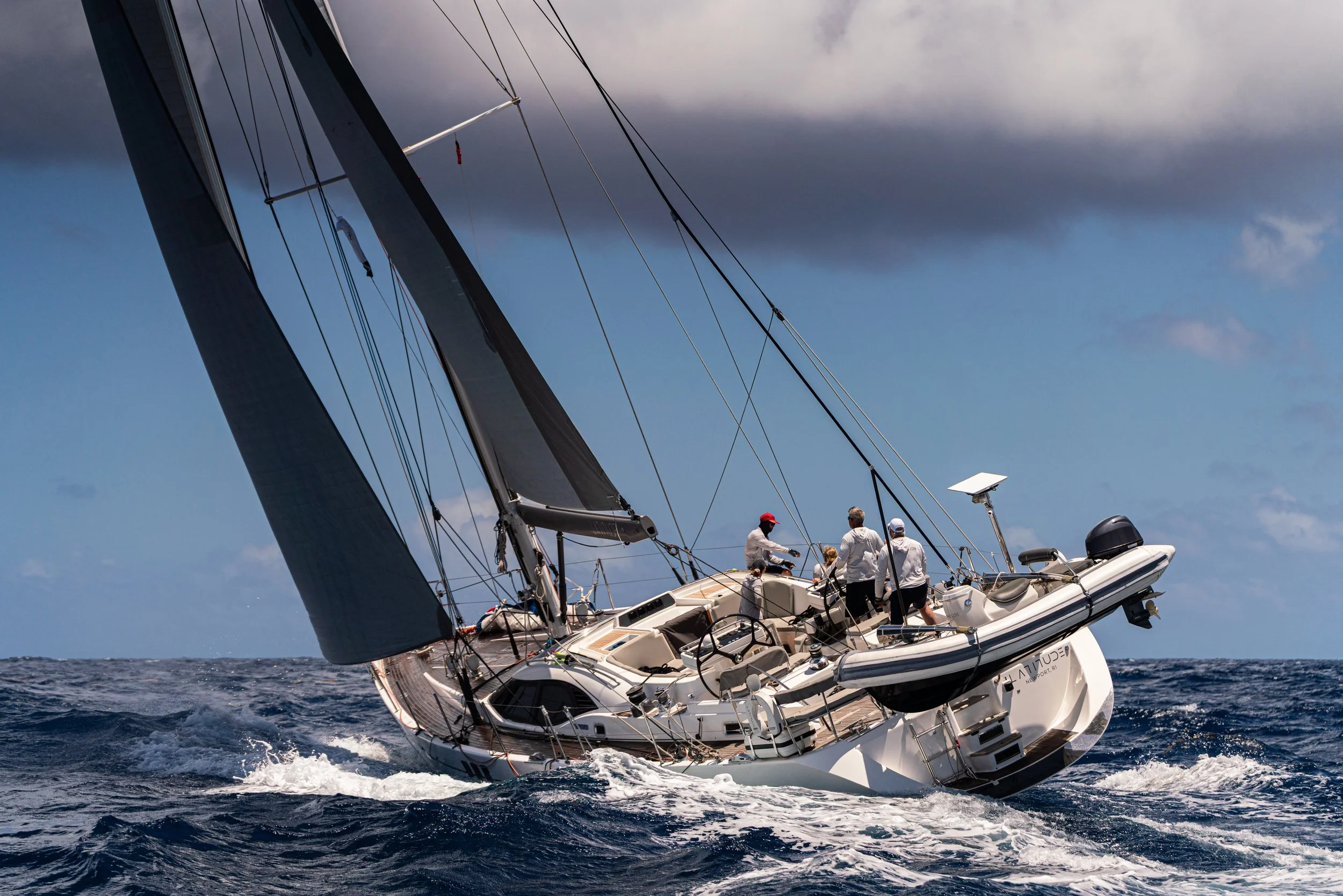 A sailing yacht tilting on the water with crew members on deck, under a partly cloudy sky.