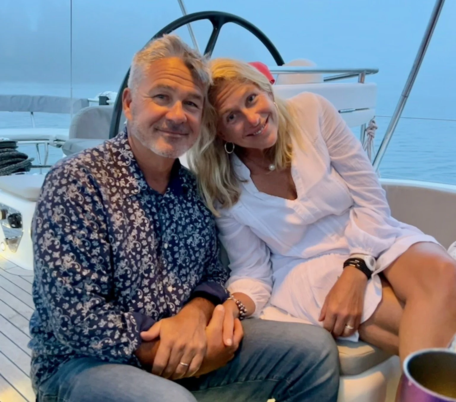 A man and woman sitting close together on a boat, smiling at the camera with a water view in the background.