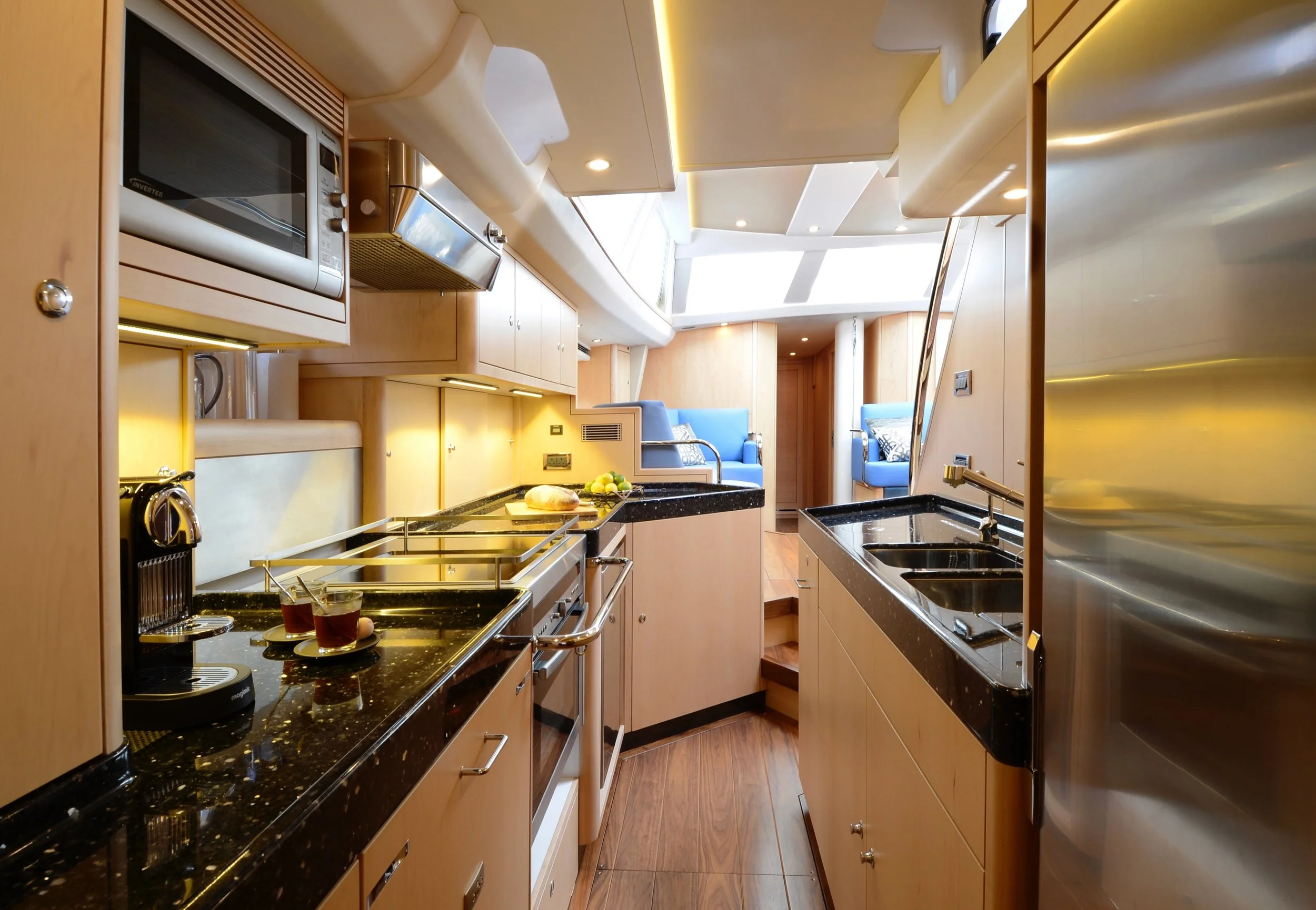 A modern kitchen with wooden cabinets, black granite countertops, a stainless steel refrigerator, and a microwave. There are two glasses of tea or coffee on the counter, with a loaf of bread and some lemons nearby. In the background, there is a seati