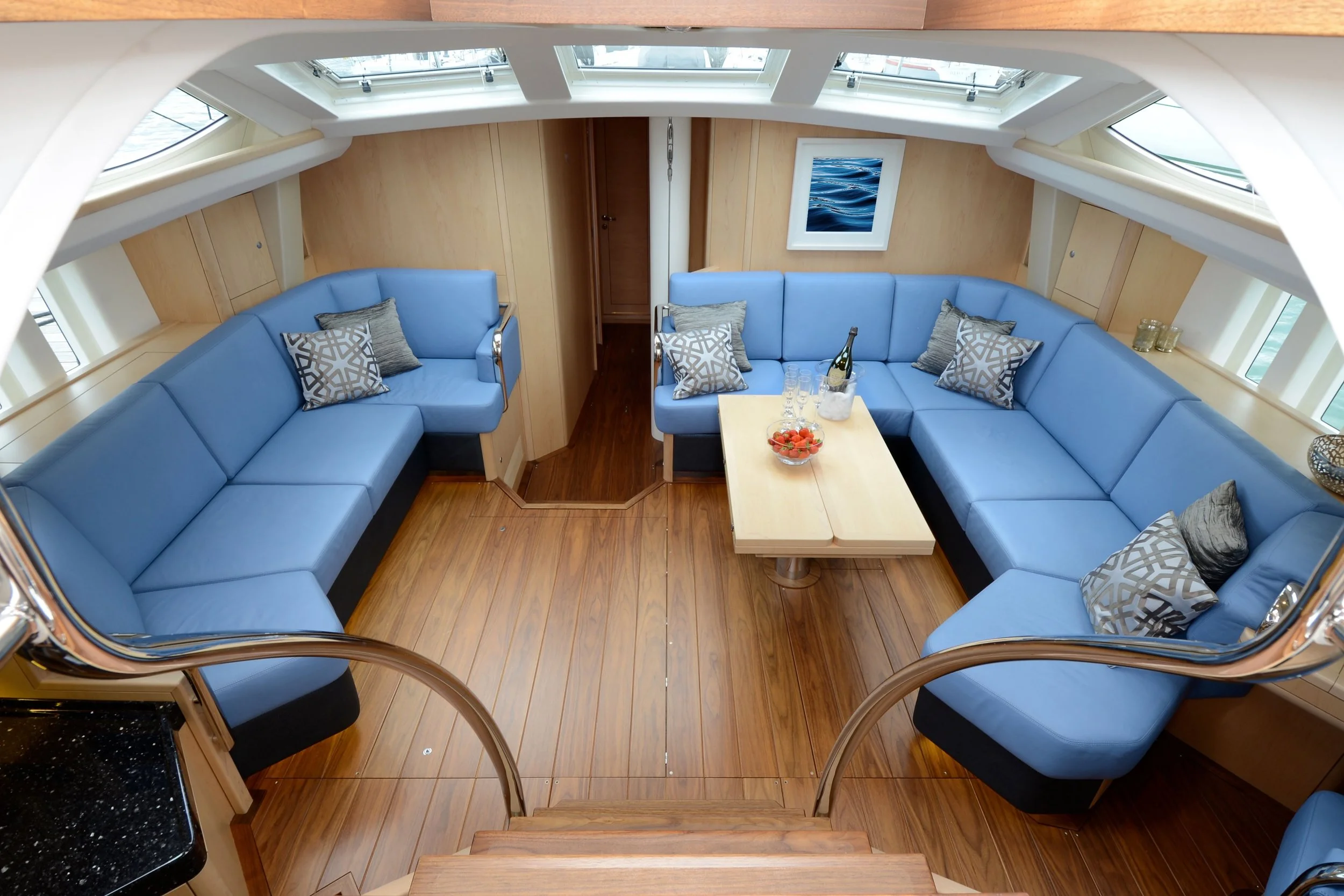 Interior of a yacht with two U-shaped blue sofas, wooden flooring, a wooden table with a bowl of strawberries, a bottle of wine, and glasses, and framed ocean art on the wall. Skylights above and windows on the sides provide natural light.