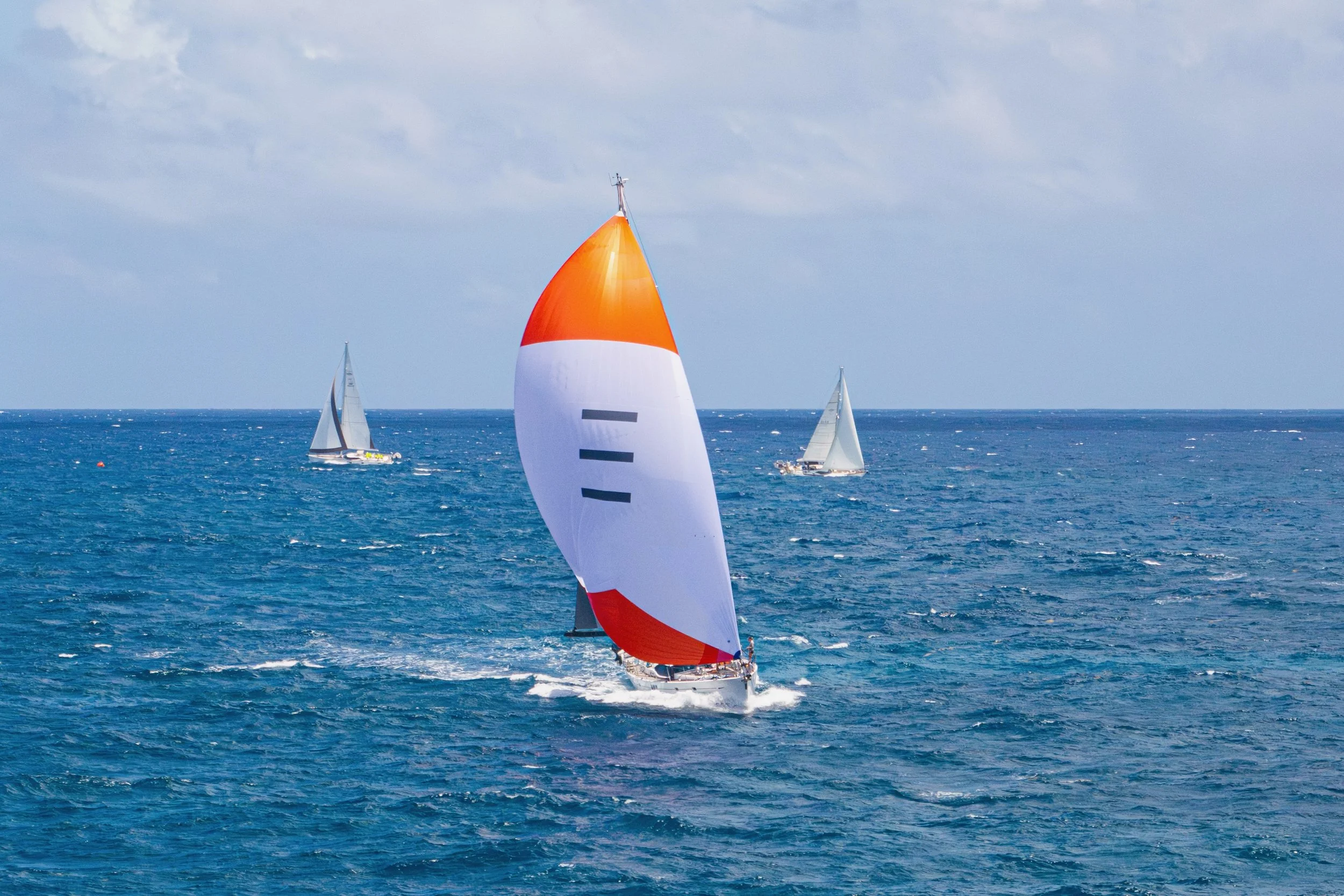 Sailing boat with a large white and orange spinnaker sail on the open ocean with two other sailboats in the background.