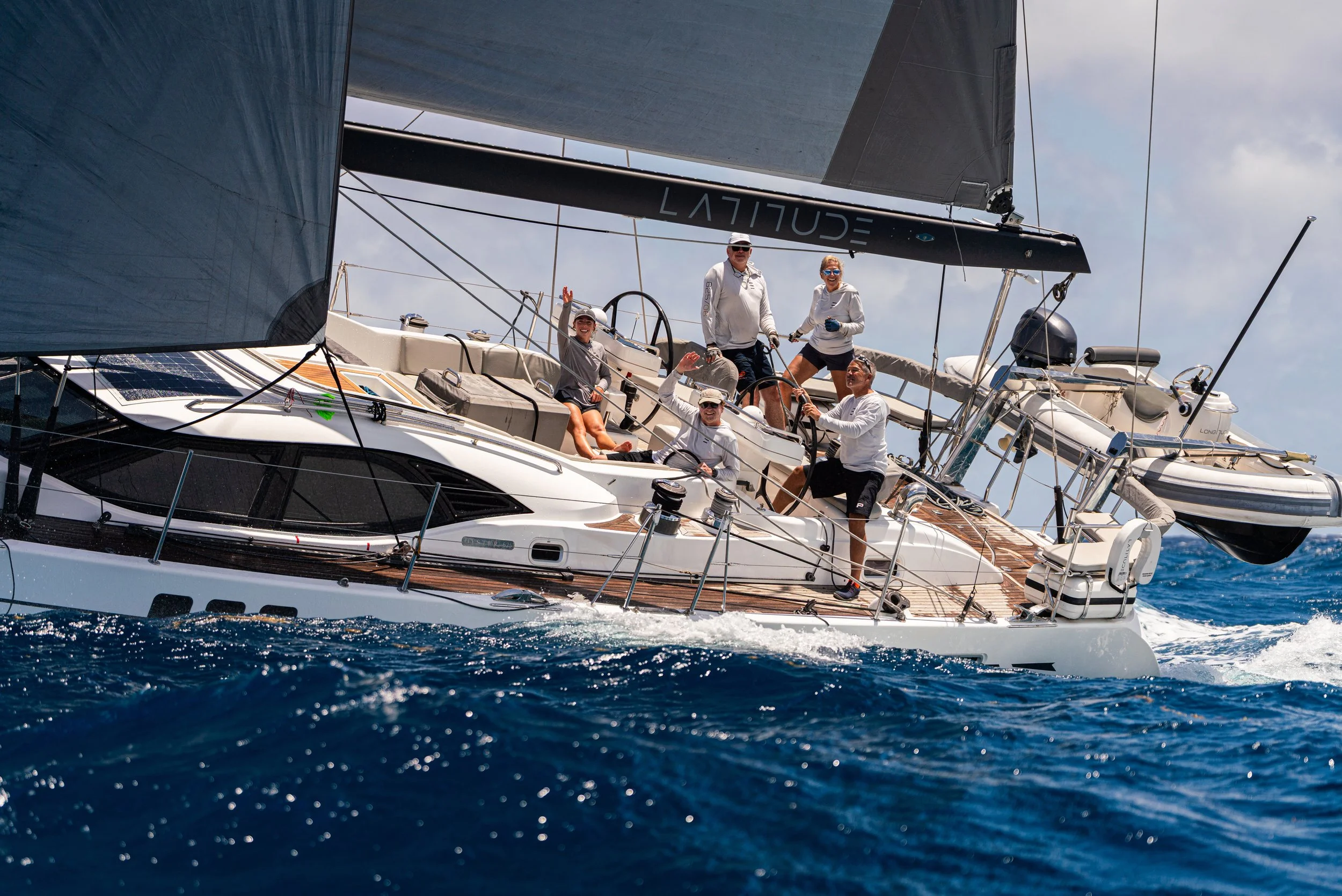 A group of seven people on a sailing yacht in open water, with a large sail, some wearing light-colored clothing and hats, enjoying a day of sailing.