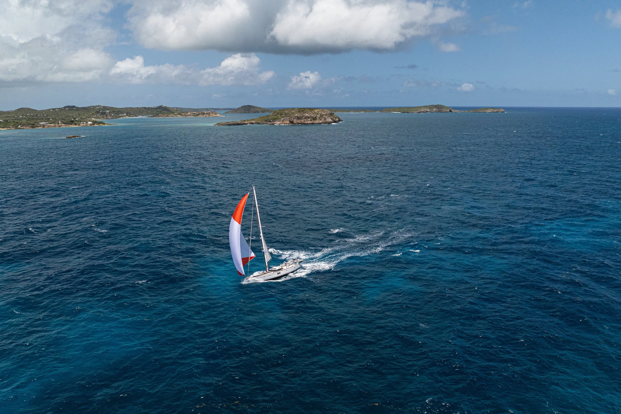 A sailboat with a red, white, and orange spinnaker sailing on deep blue ocean waters near a coastline with green land and small islands in the background under partly cloudy sky.