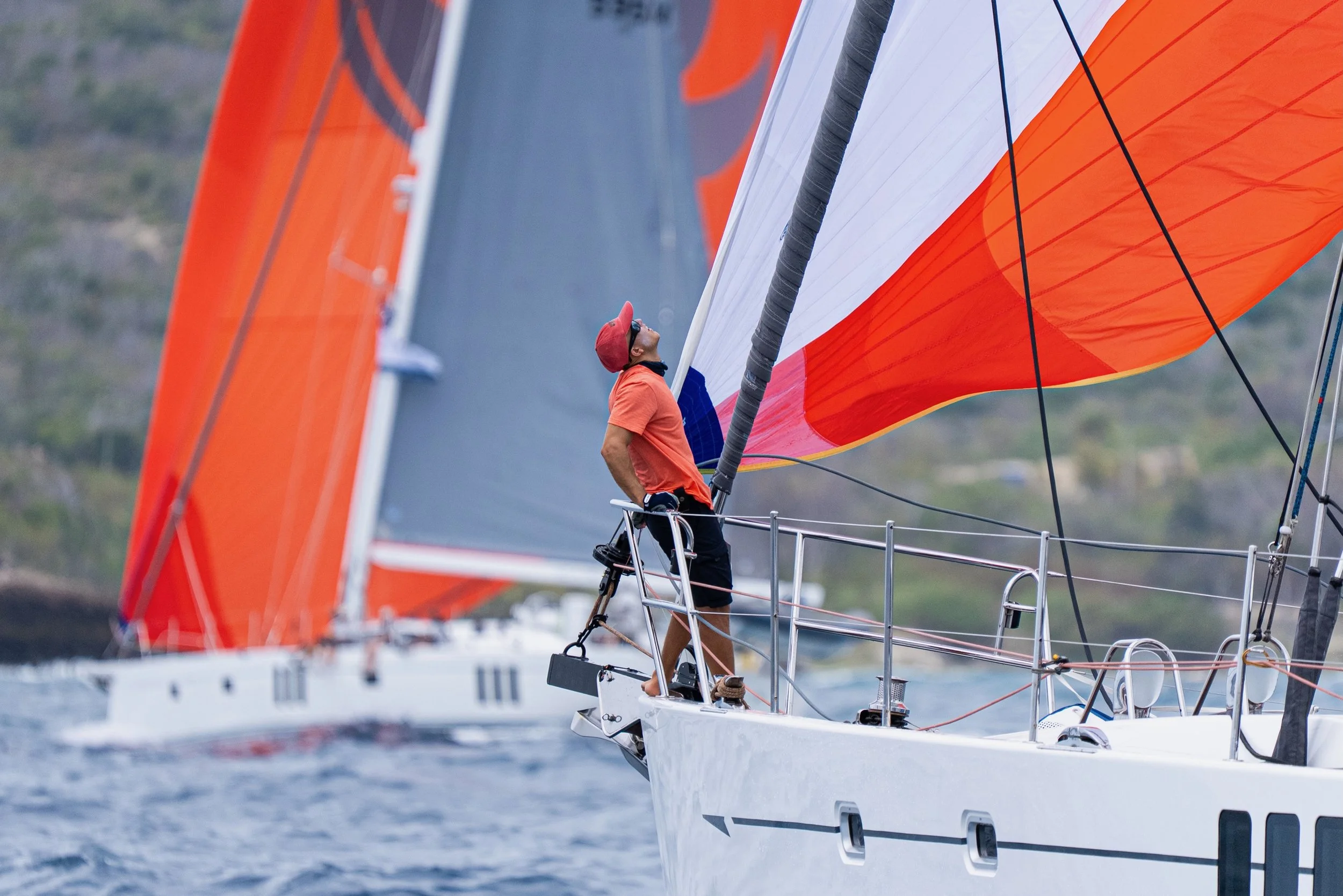 A man wearing a red cap, orange shirt, and shorts stands on the deck of a sailboat, looking up at the orange and white sails. The boat is on water with a blurred green and blue shoreline in the background.